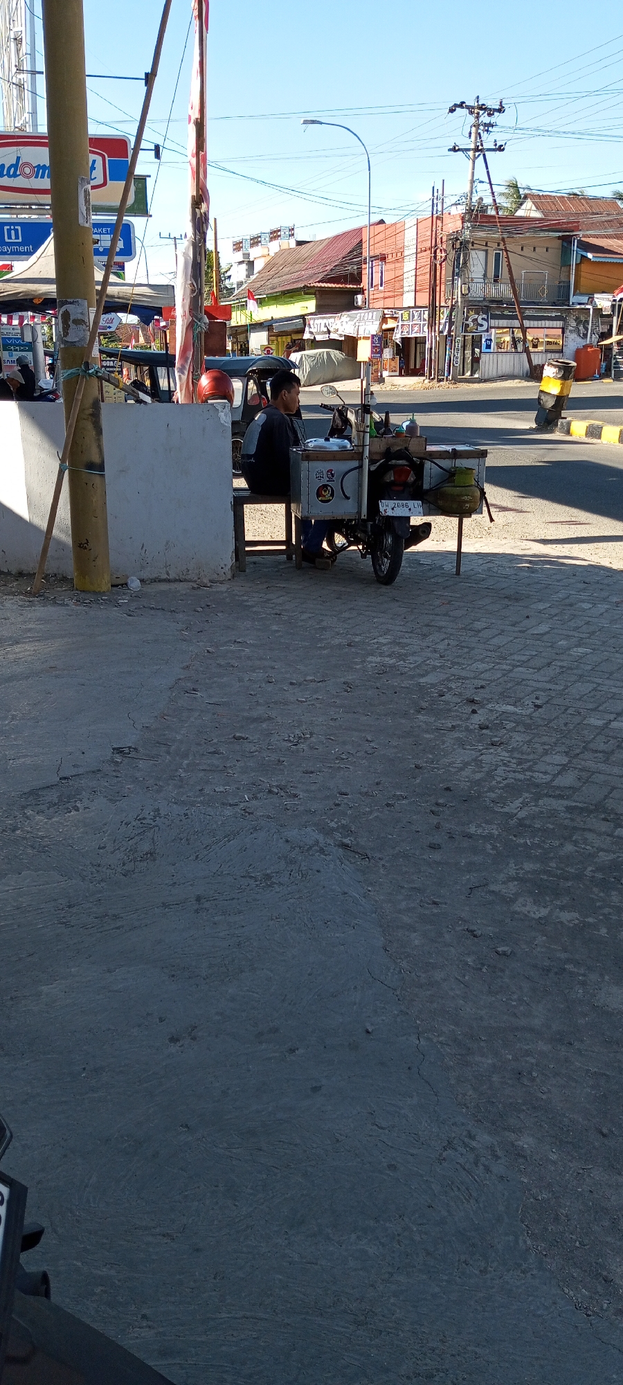 In Assorajang, Indonesia, a person is seated on a stool next to a food cart attached to a motorcycle. The cart appears to be made of metal with various containers and equipment, possibly for cooking or serving food. A green gas cylinder is visible on the side of the cart. The motorcycle has a license plate that reads "UH 2086 LH". The person is wearing a dark long-sleeved shirt and jeans. The scene is set on a street with a paved surface and a sidewalk. In the background, there are buildings, a street lamp, power lines, and a clear blue sky. The overall impression is of a street vendor operating their business in an urban or semi-urban environment.