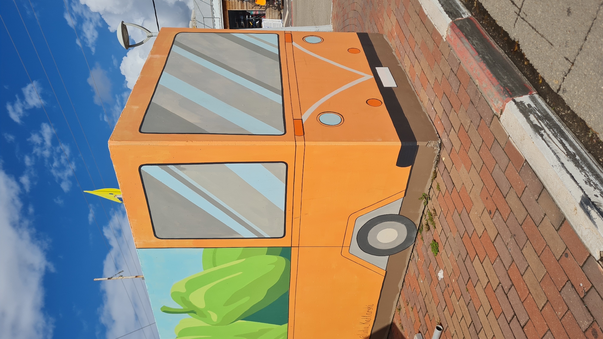 The image shows a painted, orange Volkswagen bus on a concrete block in Nahariyya, Israel. The bus has large, grey-tinted windows with blue and grey stripes. The top of the bus has orange headlights and a white license plate, separated by a curved line. One side of the bus has a painted wheel with a grey hubcap. Underneath the bus windows on one side, there are painted green bell peppers. The bus sits on a brick walkway next to a white curb. In the background is a blue sky with white clouds and power lines. A street light is visible behind the bus.