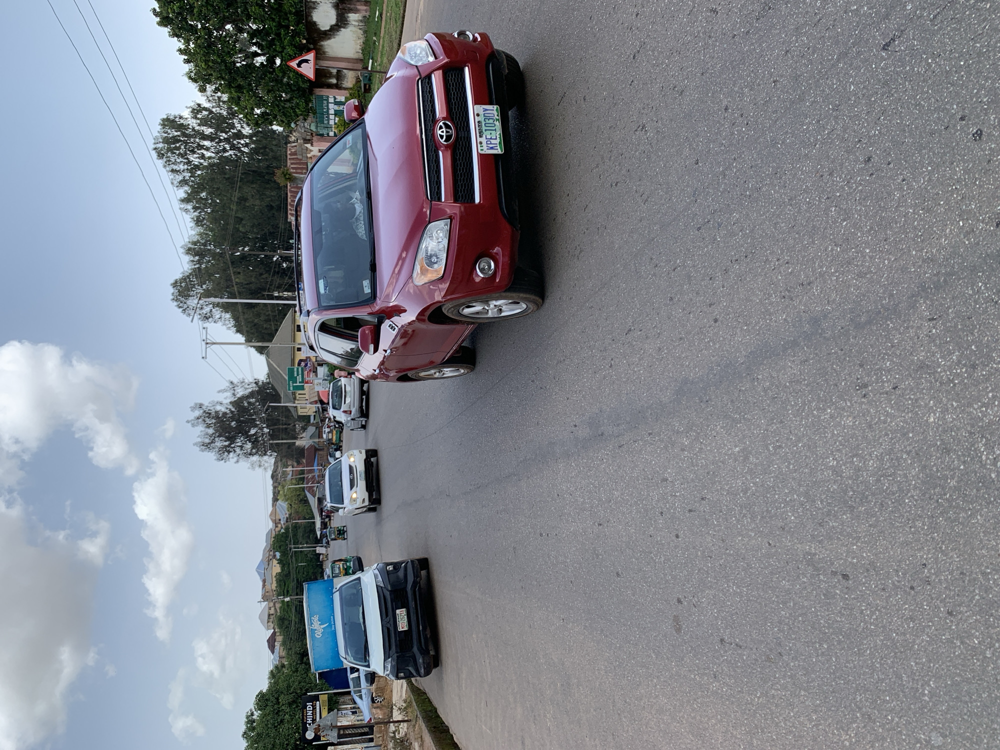 The image shows a street scene in Jos, Nigeria, with a clear view of multiple vehicles on an asphalt road. In the foreground, a red Toyota RAV4 is prominently featured, with its license plate clearly visible as "KPE-103DY". To the left and slightly behind the RAV4, a white Mitsubishi Pajero is parked or driving, displaying the license plate "MD0-2621A". Further down the road, other vehicles, including a white Lexus SUV and various other cars and a tricycle, are visible, indicating moderate traffic. The environment appears to be a commercial or residential area, with buildings, trees, and utility poles lining the street. The sky is partly cloudy, suggesting daytime. A triangular traffic sign with a black curved arrow on a white background is visible on the right side of the road. The overall scene depicts everyday urban life in Jos.