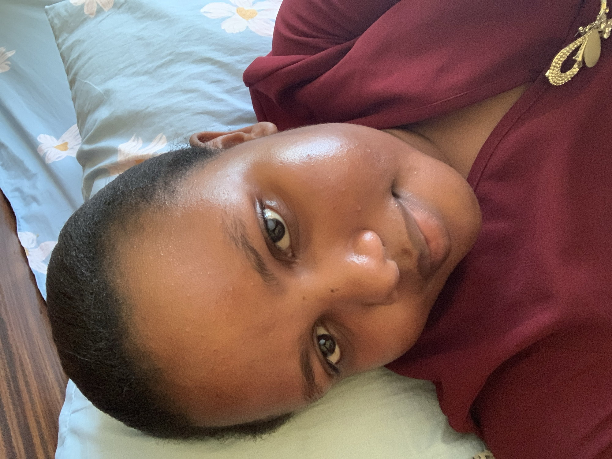 A person with dark skin and short, dark hair is lying on a light blue pillow patterned with white flowers. They are wearing a maroon-colored garment with a gold-colored brooch. The person is looking at the camera with a slight smile. The background is a wooden floor. This scene cannot be identified as specifically in Dar es-Salaam, Tanzania without additional information.