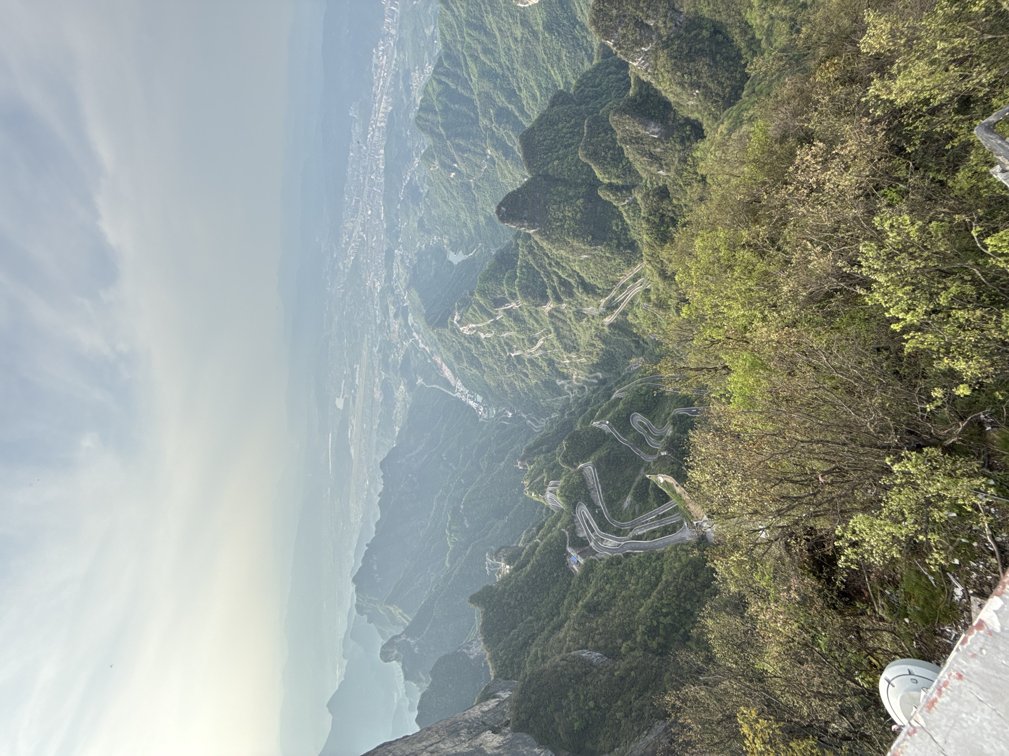 A high-angle view captures Tianmen Mountain Road, also known as the 99 turns road, descending through lush, green mountains in the Yongding District of China. The road is comprised of winding hairpin turns. In the distance, a city, possibly Zhangjiajie, is visible through the hazy atmosphere. The foreground includes trees and vegetation. At the very bottom right is a structure, possibly a walkway.