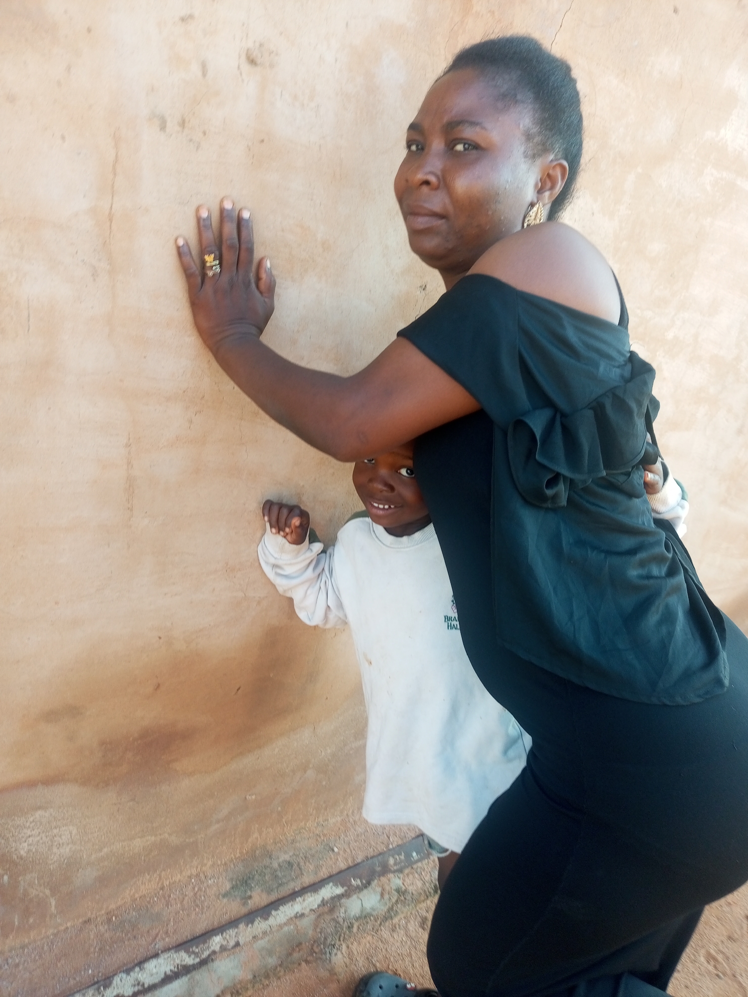 In Tempure, Nigeria, a dark-skinned woman, wearing a black off-the-shoulder top and black pants, leans against a tan, textured wall. Her left hand, adorned with rings, is pressed against the wall. Partially visible behind her and to her left is a young child in a white long-sleeved shirt, looking towards the camera. The background is an unpainted wall with visible texture and some discoloration. The lighting suggests an outdoor setting.