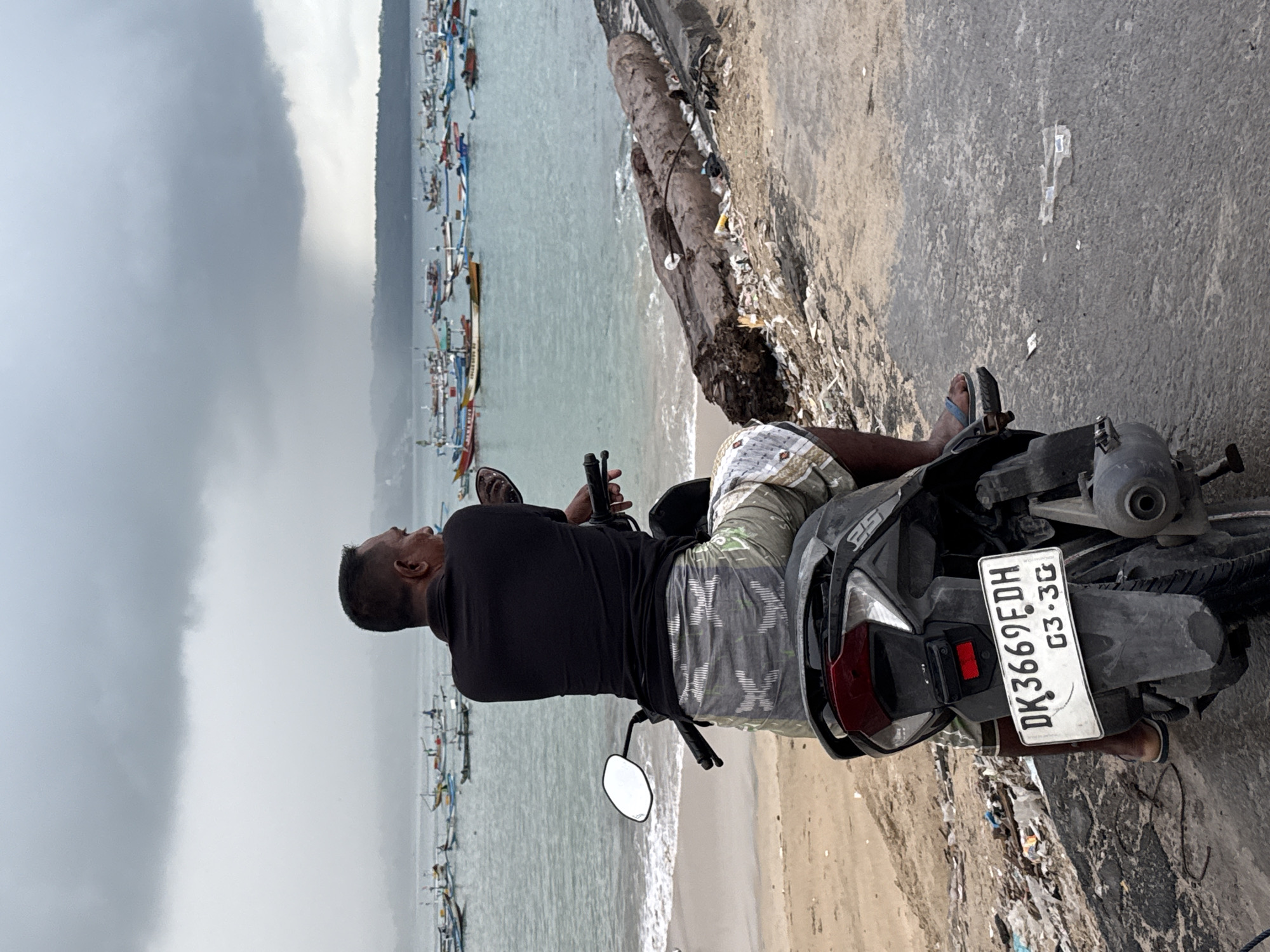 A man is depicted sitting on a dark-colored scooter, parked alongside a concrete seawall at a coastal location in Kuta, Indonesia. He is seen from behind, looking out towards the sea, with his left arm resting on the scooter's handlebar. He wears a black long-sleeved shirt, camouflage-patterned shorts, and blue flip-flops on one foot.
The scene captures a relaxed moment as the man observes the activity on the water. Numerous traditional Indonesian fishing boats, known as Jukungs, are visible in the light blue-green water, lined up along the shore. The immediate foreground shows a paved surface where the scooter is parked, while a sandy beach area with some debris, including logs, is visible between the seawall and the water. The sky is overcast and grey, suggesting a cloudy day.
The scooter's license plate is clearly visible, displaying "DK 3669 EDH" and "03.30", which is consistent with vehicle registrations in Bali, Indonesia. The overall atmosphere is quiet and contemplative.