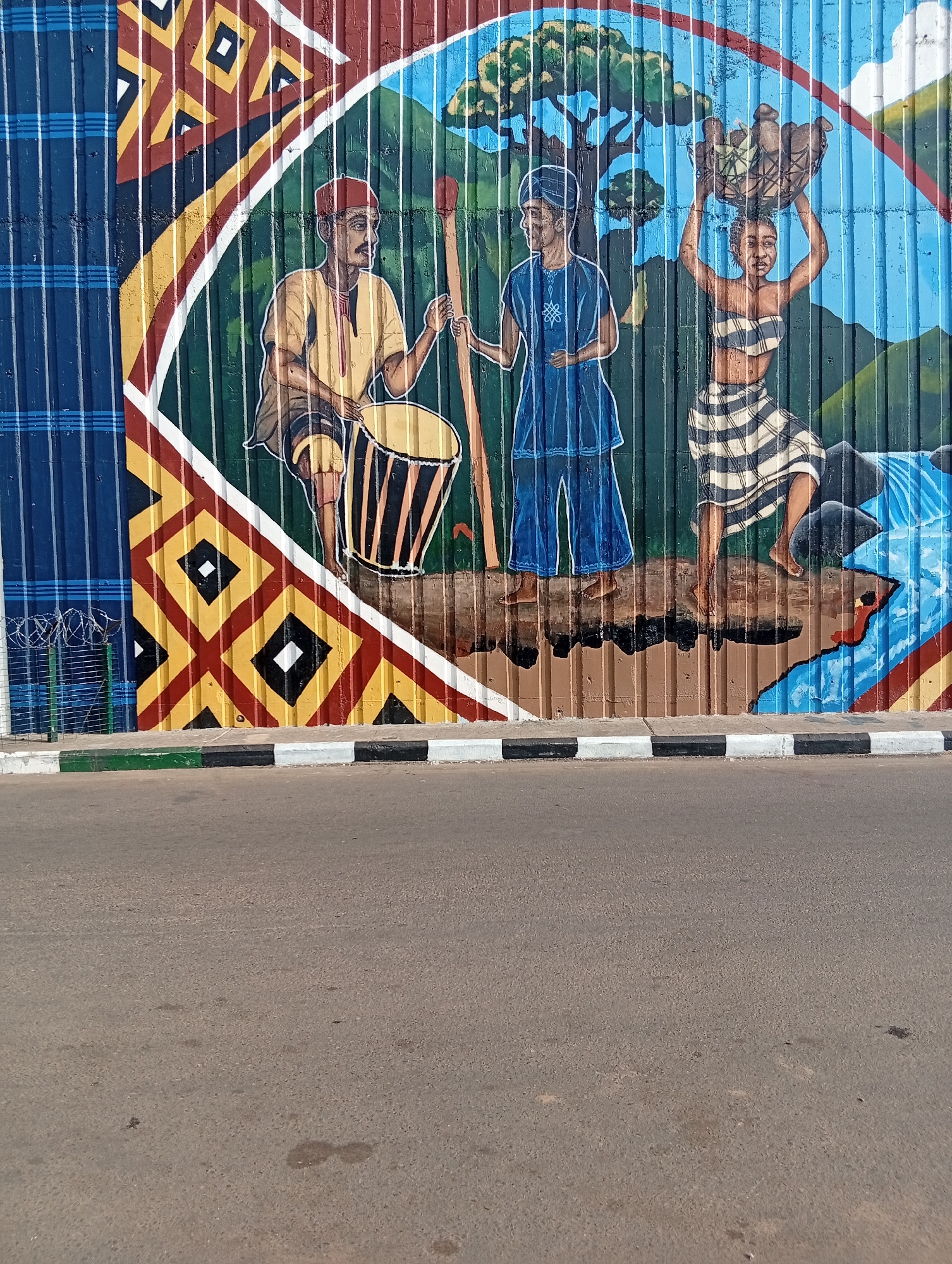 The image shows a colorful mural painted on a corrugated metal wall in Jalingo, Nigeria. The mural depicts three people in traditional attire. On the left, a man wearing a red cap and a yellow tunic is sitting and playing a drum. Next to him, a person in a blue robe stands holding a long wooden staff. On the right, a woman with her hair tied up is carrying a basket of produce on her head, wearing a striped top and skirt. Behind the figures, the mural features a landscape with trees, mountains, and a waterfall. The bottom of the mural is bordered by a decorative pattern of diamonds and squares in yellow, red, and black. In front of the mural is a paved road with a curb painted with black and white stripes, and a small patch of grass with a fence made of wire mesh and barbed wire. The overall scene suggests a depiction of traditional life and culture in Nigeria.
