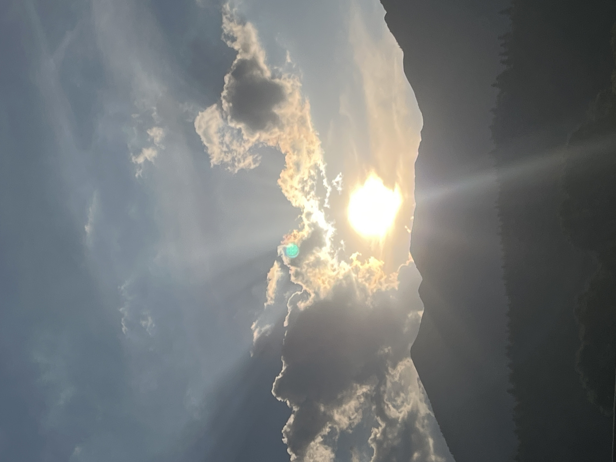 This is a vertical, slightly low-angle shot of a dramatic sky over Garmisch-Partenkirchen, Germany. The sun is a bright, intense orb, partially obscured by a large, dark cloud formation that casts a sharp shadow on the right side of the frame. Rays of light pierce through the clouds, creating a dramatic effect. The clouds themselves are varied, with some dark and ominous, while others are illuminated by the sun, appearing white and wispy. In the background, on the right side, a dark, silhouetted mountain range is visible, covered in trees. The sky transitions from a bright, sunlit area on the left to a darker, hazy atmosphere on the right. The overall impression is one of a powerful natural display, possibly during a storm or a dramatic sunset/sunrise. The light suggests it is either early morning or late afternoon. No people, objects, or text are visible in the image.