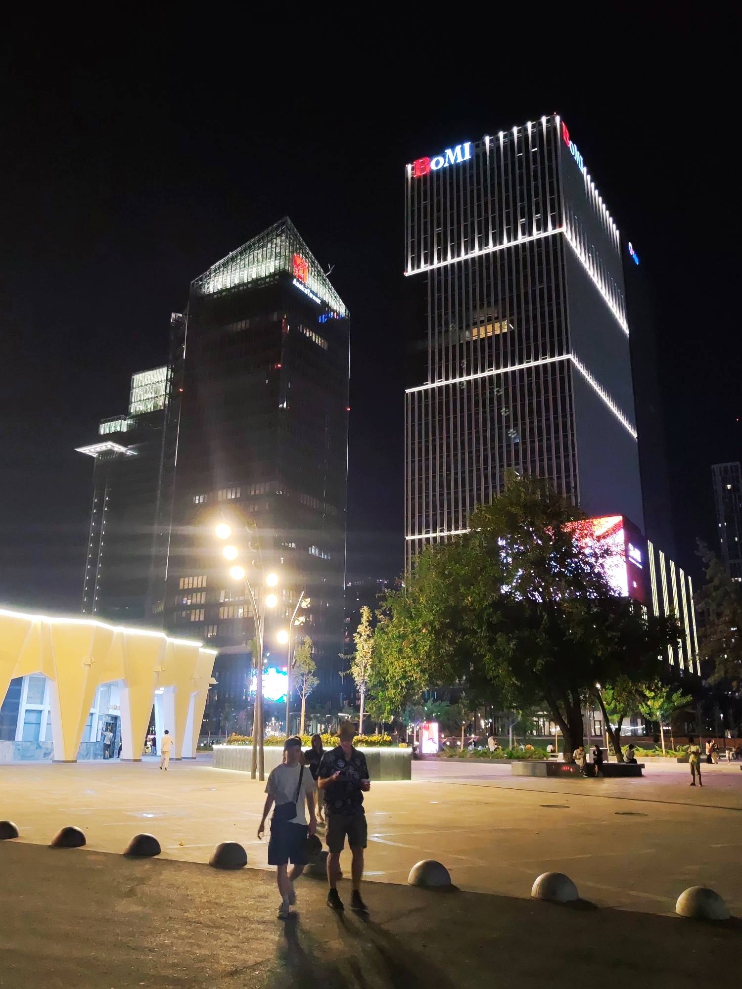 The image depicts a nighttime scene in Tashkent, Uzbekistan, likely in the vicinity of the City Park or a similar modern urban development. Two prominent, modern skyscrapers dominate the background. The taller building on the right is illuminated with white vertical lights and displays the logo "BOMI" in red and blue lettering at its summit. The building on the left has a more angular design with green illumination on its upper section and a red logo near its peak. In the mid-ground, a contemporary covered walkway with yellow, illuminated geometric supports is visible. Several streetlights with multiple bright bulbs are positioned along a pathway. A tree with lush green foliage stands between the two skyscrapers. In the foreground, a paved plaza area is populated by several individuals. Two young men are walking towards the viewer; one in a light-colored t-shirt and dark shorts, wearing a crossbody bag, and the other in a dark patterned shirt and shorts. Further in the background, various people are seen seated or walking in the plaza and park areas, with more trees and illuminated structures suggesting a public gathering space. The overall atmosphere is one of a well-lit, modern urban environment at night.