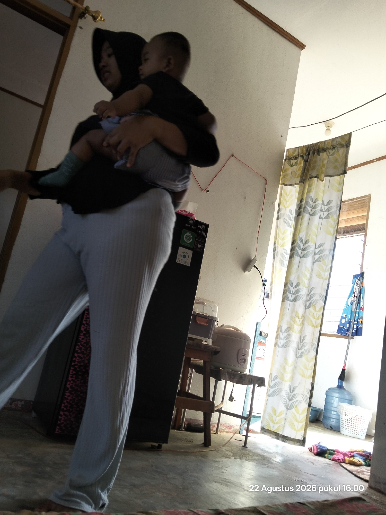 A full-body, indoor, medium-shot photograph captures a person carrying a baby. The person, wearing a black hijab and light grey ribbed pants, is positioned on the left side of the frame, facing towards the right. The baby, wearing a black shirt and light blue and grey striped pants, is held in the person's arms, facing away from the camera. The background includes a dark refrigerator with stickers, a rice cooker on a wooden stand, a partially drawn curtain with a leaf pattern, and a large blue water jug. The floor appears to be concrete. A timestamp in the lower right corner reads "22 Agustus 2026 pukul 16.00".