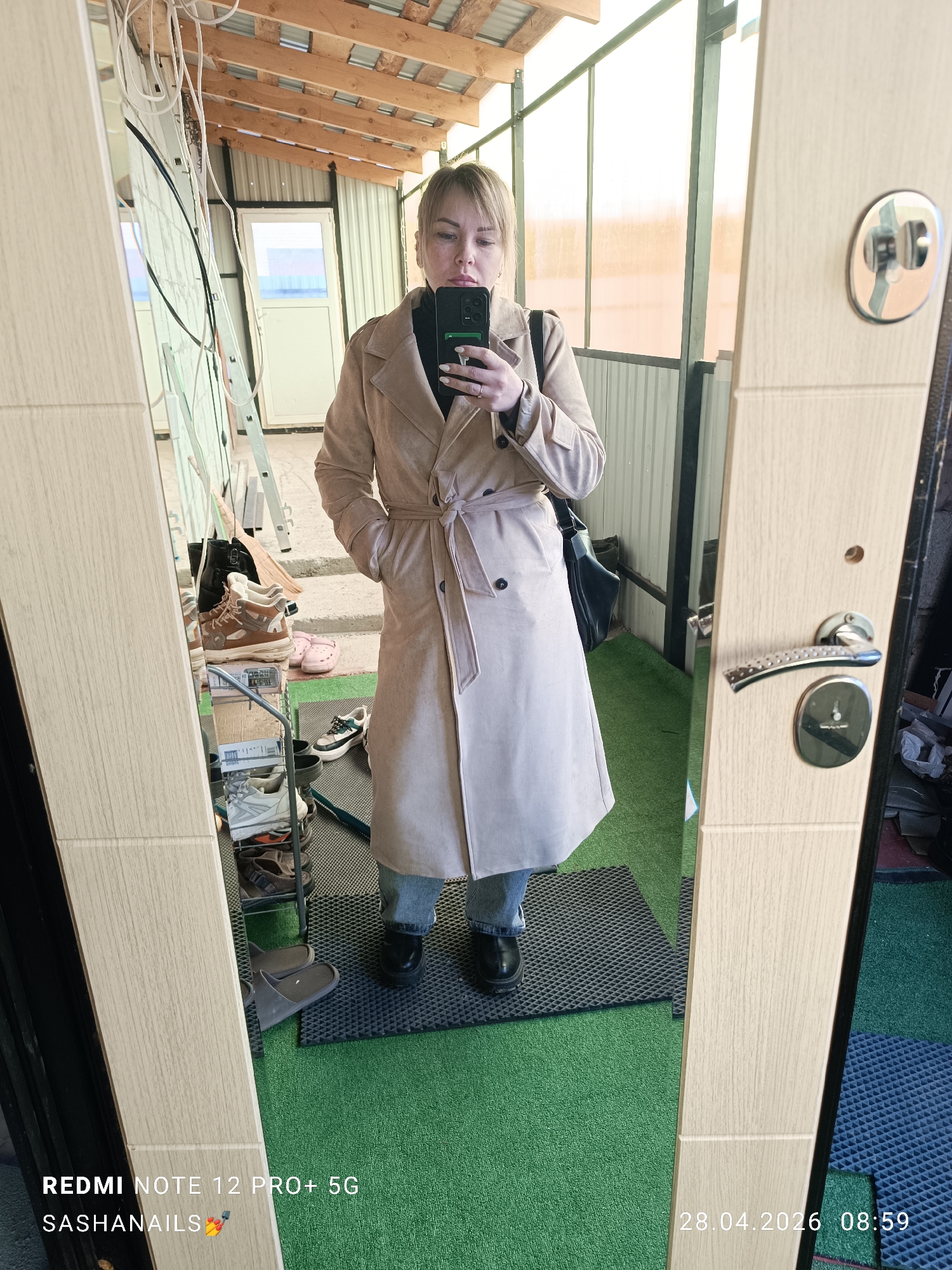 A woman is captured in a mirror selfie taken in what appears to be an enclosed porch or sunroom in Chaikovsky, Russia. The image was taken at 08:59 on April 28, 2026. She has light-colored hair, a neutral expression, and is wearing a light beige trench coat tied at the waist, a black turtleneck, blue jeans, and black chunky-soled shoes. She holds a dark smartphone, which is visible in her left hand. A black bag is slung over her left shoulder.
The setting features an informal structure with wooden ceiling beams, translucent corrugated roofing, and corrugated metal walls. The floor is covered with green artificial grass and a grey textured mat. Reflected in the mirror to her left, a shoe rack displays several pairs of sneakers and sandals, alongside a ladder. The right side of the mirror shows part of a light-colored wooden door frame with a door handle and lock. Bright natural light illuminates the space through the translucent roof and a white door/window in the background.
Visible text includes "REDMI NOTE 12 PRO+ 5G" and "SASHANAILS π
β¨" at the bottom left, and the timestamp "28.04.2026 08:59" at the bottom right.
