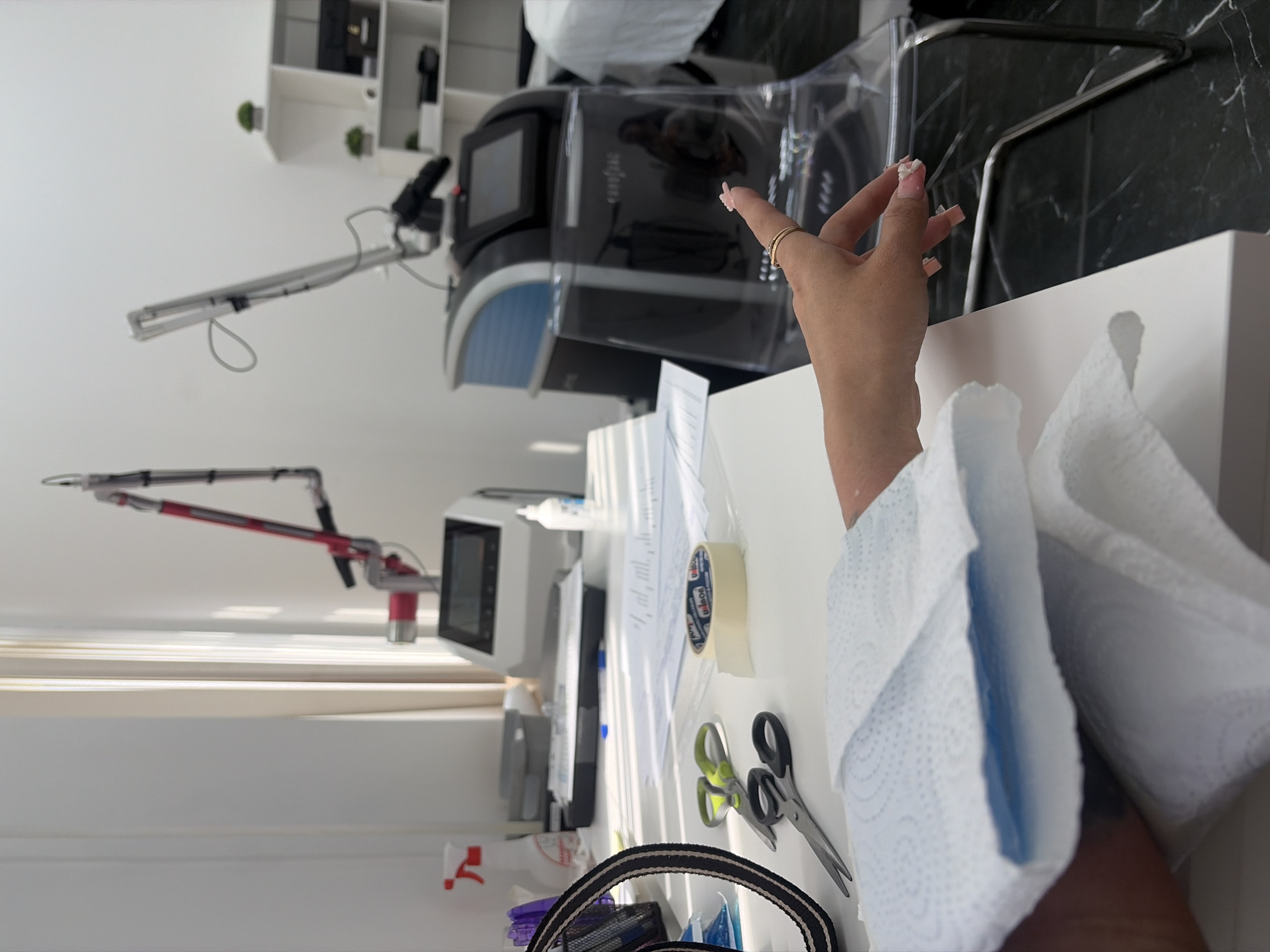 An indoor scene, likely a medical or aesthetic treatment room, is depicted with medical equipment and a person's arm. The room features a white desk with various items, including a red spray bottle, scissors, masking tape, and papers. Two laser treatment machines are visible in the background, one red and silver, the other black and blue. A person's left arm, covered with blue and white paper towels, rests on the desk. The individual's right hand, adorned with rings and long, pink-tipped nails, is reaching out, with one finger pointing towards the laser equipment. The room has white walls and a window with light-colored curtains. The floor is black and white marble. The context suggests a laser treatment session in progress or preparation for one. The location is identified as Saint Petersburg, Russia.