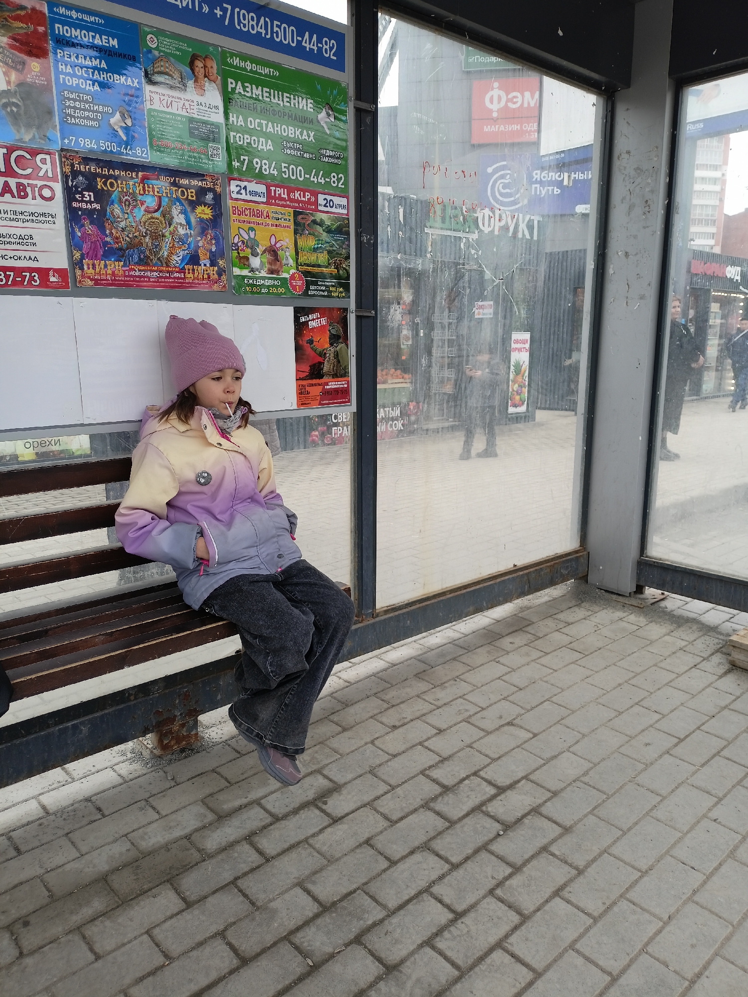 A young girl wearing a purple hat and jacket sits on a wooden bench at a bus stop in Novosibirsk, Russia. She has a lollipop in her mouth and is looking off to the side. The bus stop has glass walls and is covered in posters advertising various businesses and events, including a circus, a rabbit exhibition, and travel packages to China. The posters also list local phone numbers. Through the glass, a fruit market is visible, along with people walking by. The ground is paved with gray bricks.