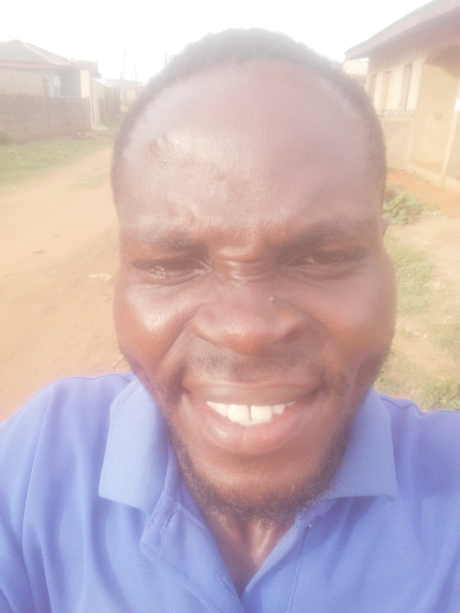 A close-up selfie shows a Black man with a dark beard and a blue collared shirt. He is smiling broadly, revealing his teeth. The background shows a dirt road and buildings under bright sunlight, suggesting an outdoor environment. The location is in the city of Unknown, Unknown.