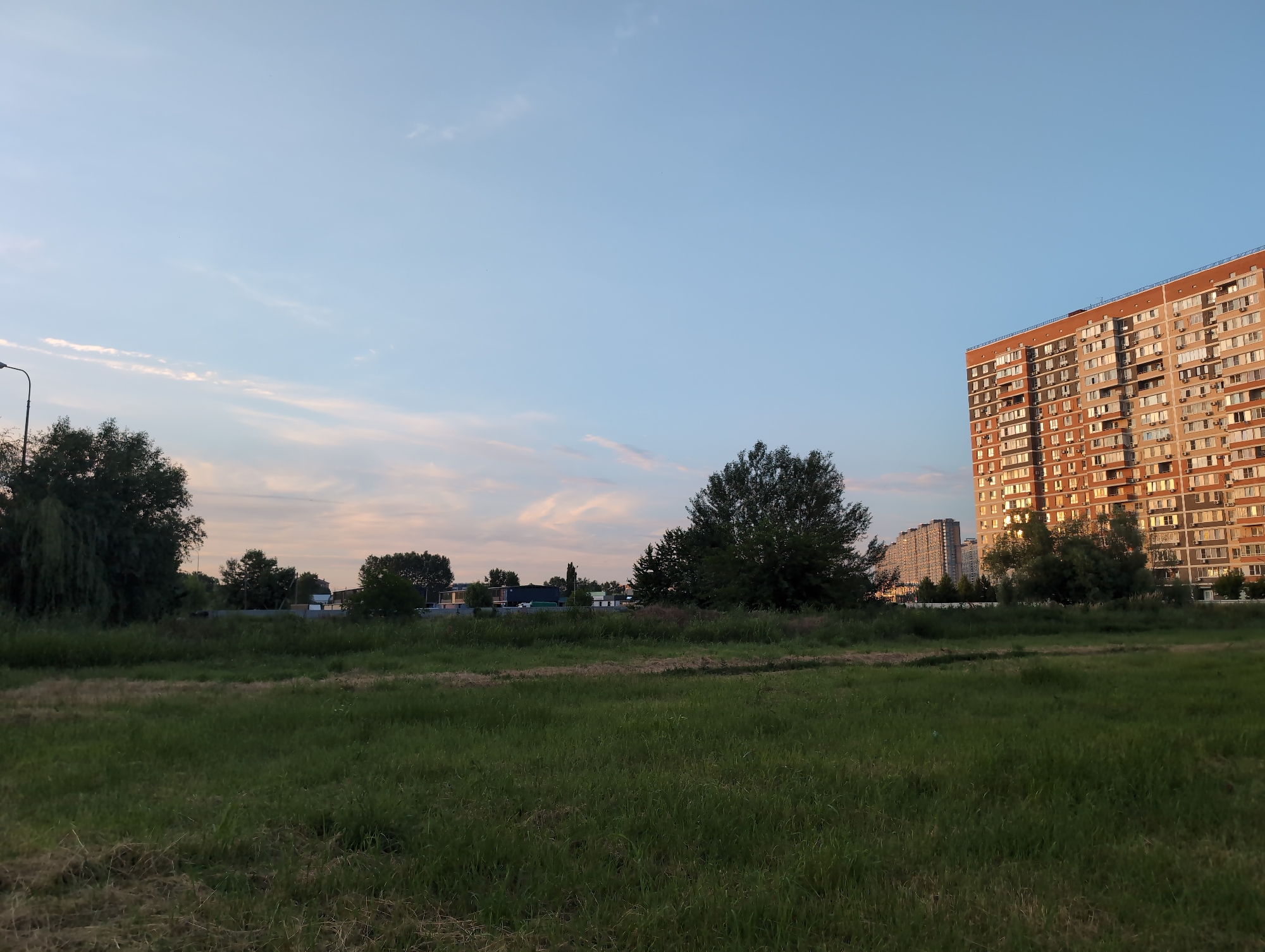 This image depicts a suburban or city edge landscape in Krasnodar, Russia, under a clear, pale blue sky with wispy clouds. The scene is dominated by a large, multi-story brick building with many windows, bathed in the warm, golden light of late afternoon or early evening. To the left of the building, a field of green grass stretches towards the foreground, with scattered trees and foliage. In the middle ground, there are more trees and some low-lying buildings or structures, possibly part of a commercial area or a different residential complex. A street lamp is visible on the far left, suggesting a developed area. The overall atmosphere is peaceful and serene, with the setting sun casting long shadows and highlighting the textures of the buildings and vegetation. There is no visible text in the image.
