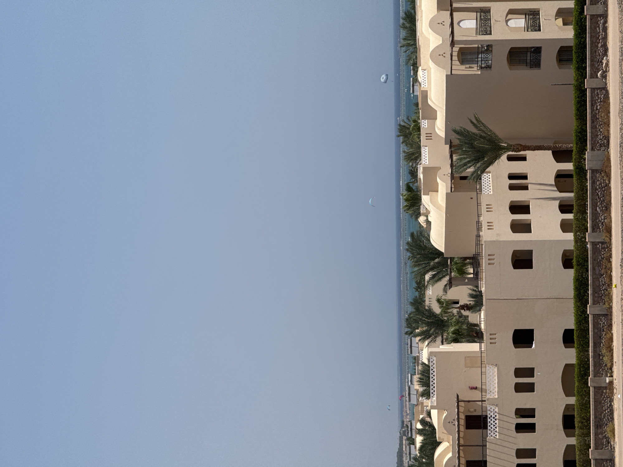 This is a wide shot of a resort with beige buildings and palm trees, likely in a coastal area of Egypt, given the characteristic architecture and clear blue sky. The resort features multiple buildings with arched windows and balconies, some with decorative white trim. Palm trees are interspersed throughout the grounds, some tall and slender, others more bushy. In the distance, the turquoise sea stretches to the horizon, with a pier or structure extending into the water. There are a few paragliders or kite surfers visible in the sky over the water, suggesting recreational activities. The foreground shows a low stone wall and a dense green hedge. The overall scene conveys a sense of a sunny, tropical vacation destination.