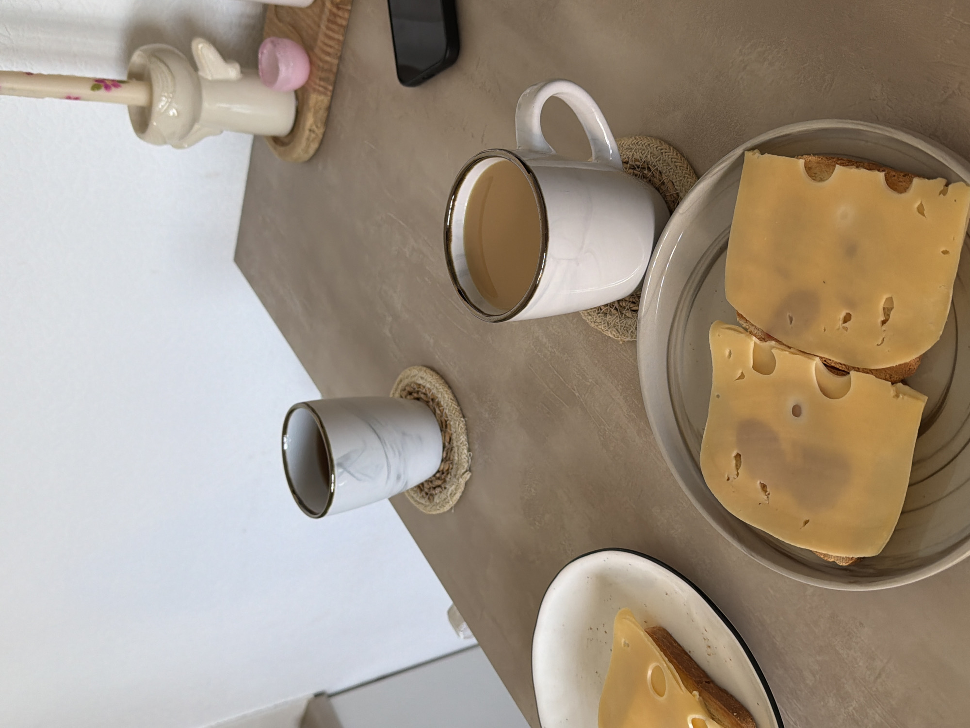 A close-up, overhead shot captures a breakfast scene on a speckled, beige countertop. In the foreground, two plates hold open-faced sandwiches topped with large slices of yellow cheese. The sandwich on the right is on a gray plate, while the one on the left is on a white plate with a black rim.
To the right of the sandwiches, a white ceramic mug with a dark rim sits on a woven coaster, filled with a light brown beverage, likely coffee or tea. Further back and to the left, another mug, this one with a marble pattern and a dark rim, rests on a similar woven coaster.
In the upper right corner, a decorative candle holder in the shape of an angel holds a pastel pink candle adorned with small floral patterns. A small pink dish sits beside it. A black smartphone is visible on the countertop near the angel figurine. The background is a plain, white wall. The overall impression is a quiet, indoor breakfast setting, with no specific indication of the city of Kyiv, Ukraine.
