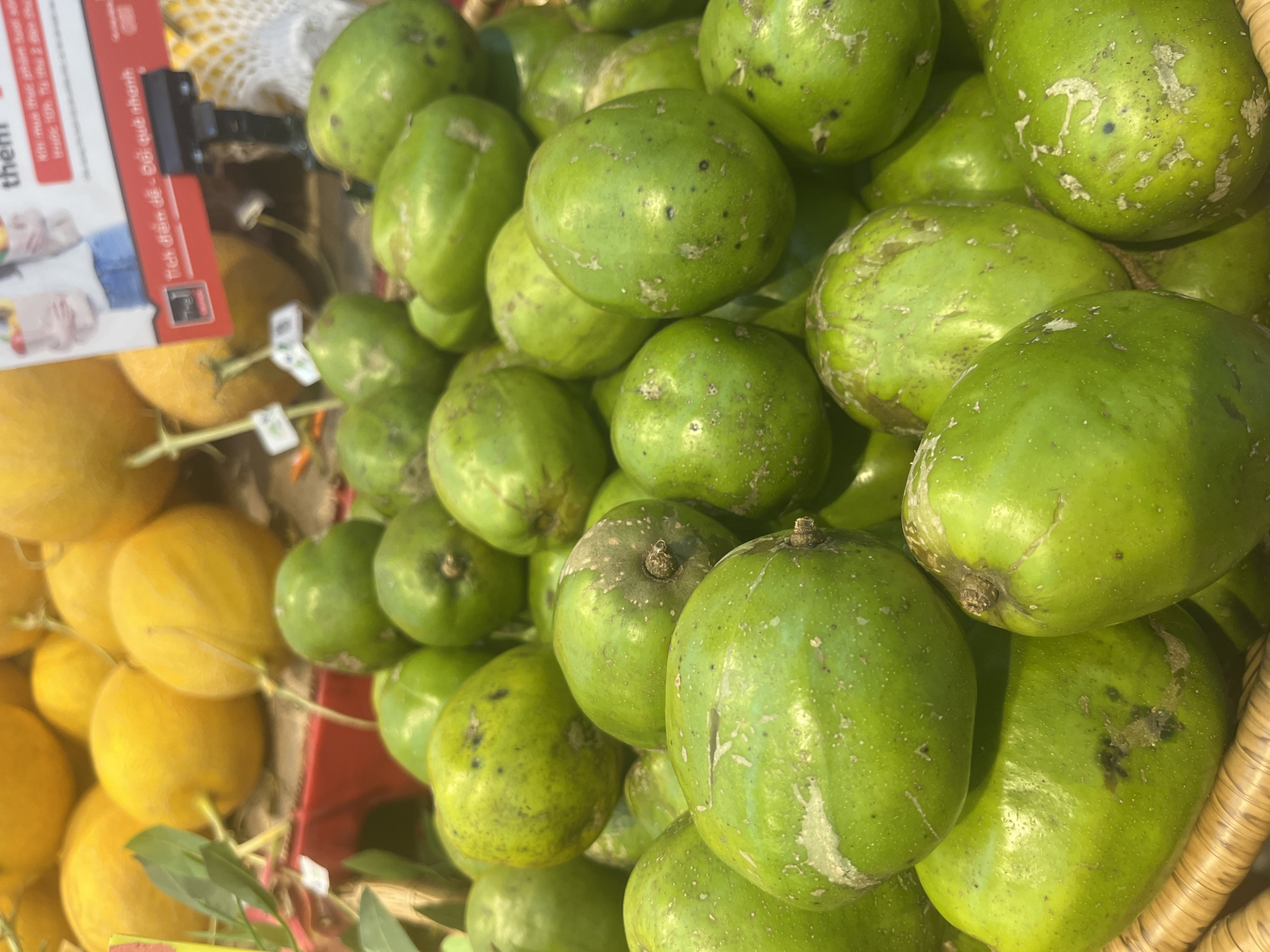 The image shows a close-up of a basket filled with green, oval-shaped fruits, likely olives or a similar fruit, with some exhibiting lighter green or white patches and darker spots. The fruits are piled high in a woven basket. In the background, there are out-of-focus yellow melons or gourds and a red bin. A portion of a sign with Vietnamese text and an image of people holding shopping bags is visible in the upper right. The lighting suggests an indoor environment, possibly a market or grocery store. The location is identified as Unknown, Unknown.