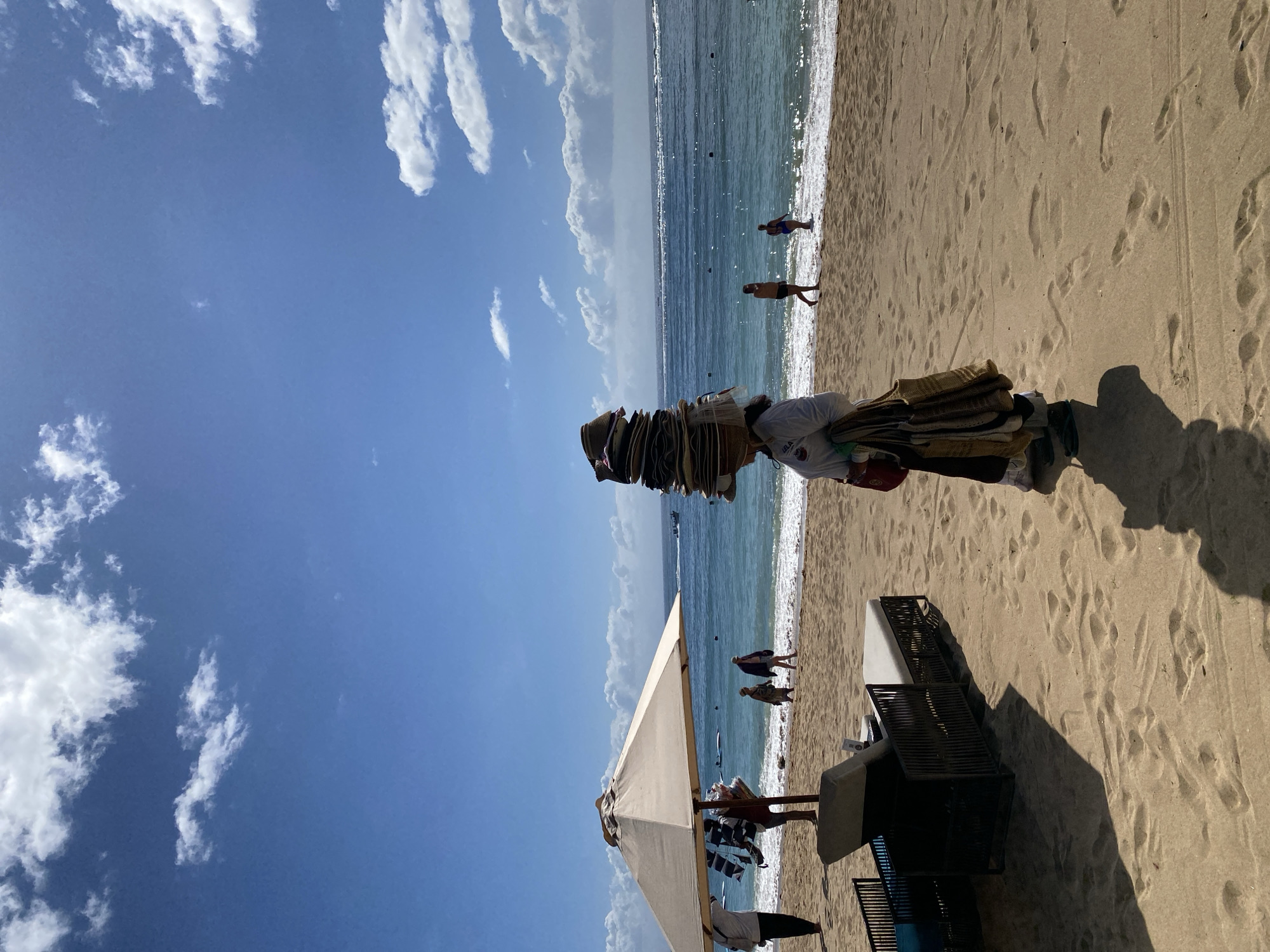 The image depicts a beach scene in Kuta Selatan, Indonesia, on a sunny day. The sky is blue with scattered white clouds. The beach is sandy with visible footprints. The ocean is turquoise, with gentle waves.
In the foreground, a vendor carries a tall stack of hats on their head. Further down the beach, several people are walking along the shoreline. An umbrella and lounge chairs are also present.