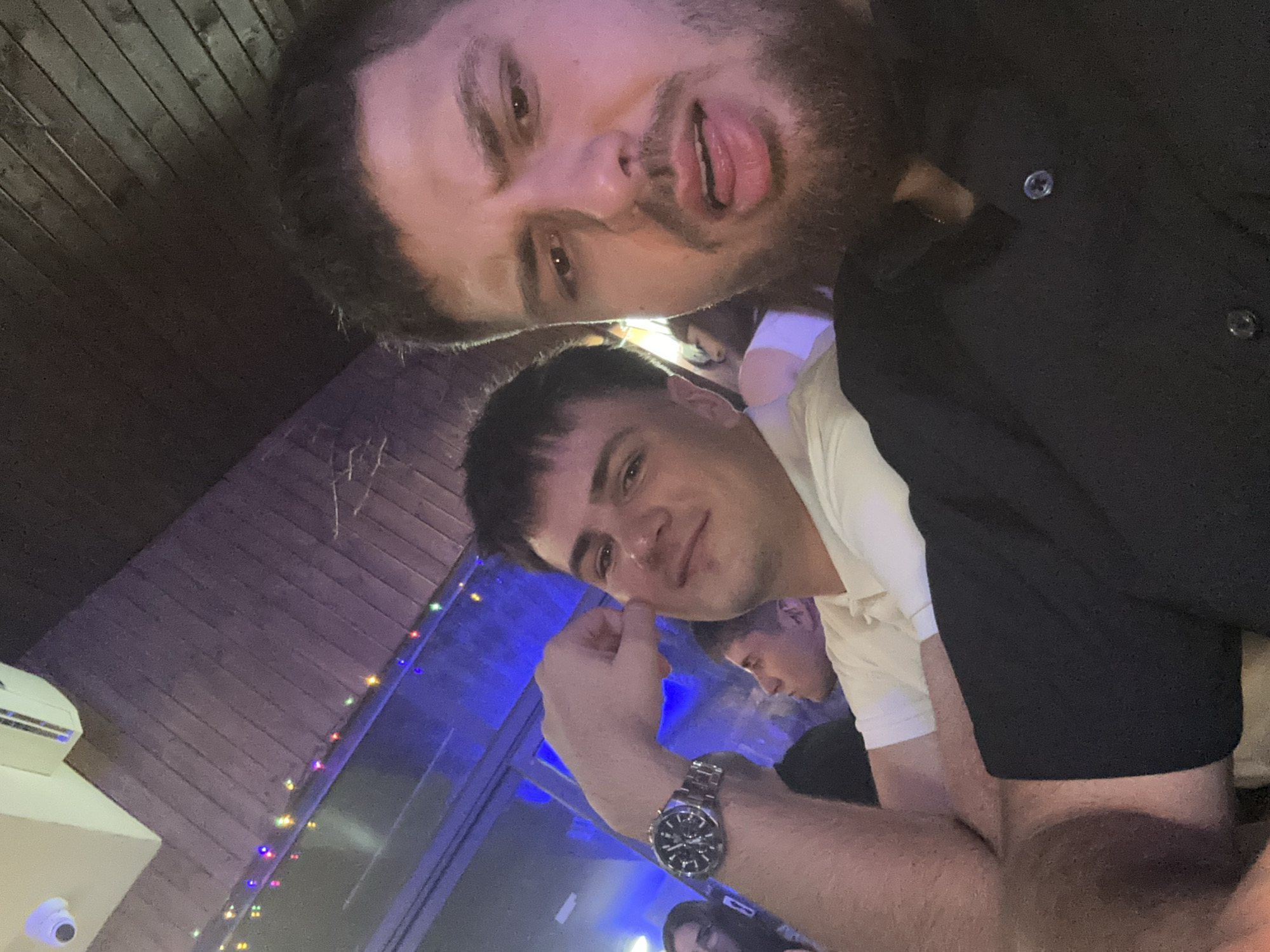 The image shows a selfie of two young men, possibly in a bar or club setting. The man in the foreground is wearing a white collared shirt and a watch on his left wrist. Behind him, another man is partially visible, appearing to wear a black shirt. The man at the top of the image is sticking his tongue out. The background shows dimly lit surroundings with colored lights and what appears to be a wooden ceiling. A security camera is visible on the top left of the image. It is not possible to determine if the location is Panagyurski kolonii, Bulgaria, from the visual information provided.