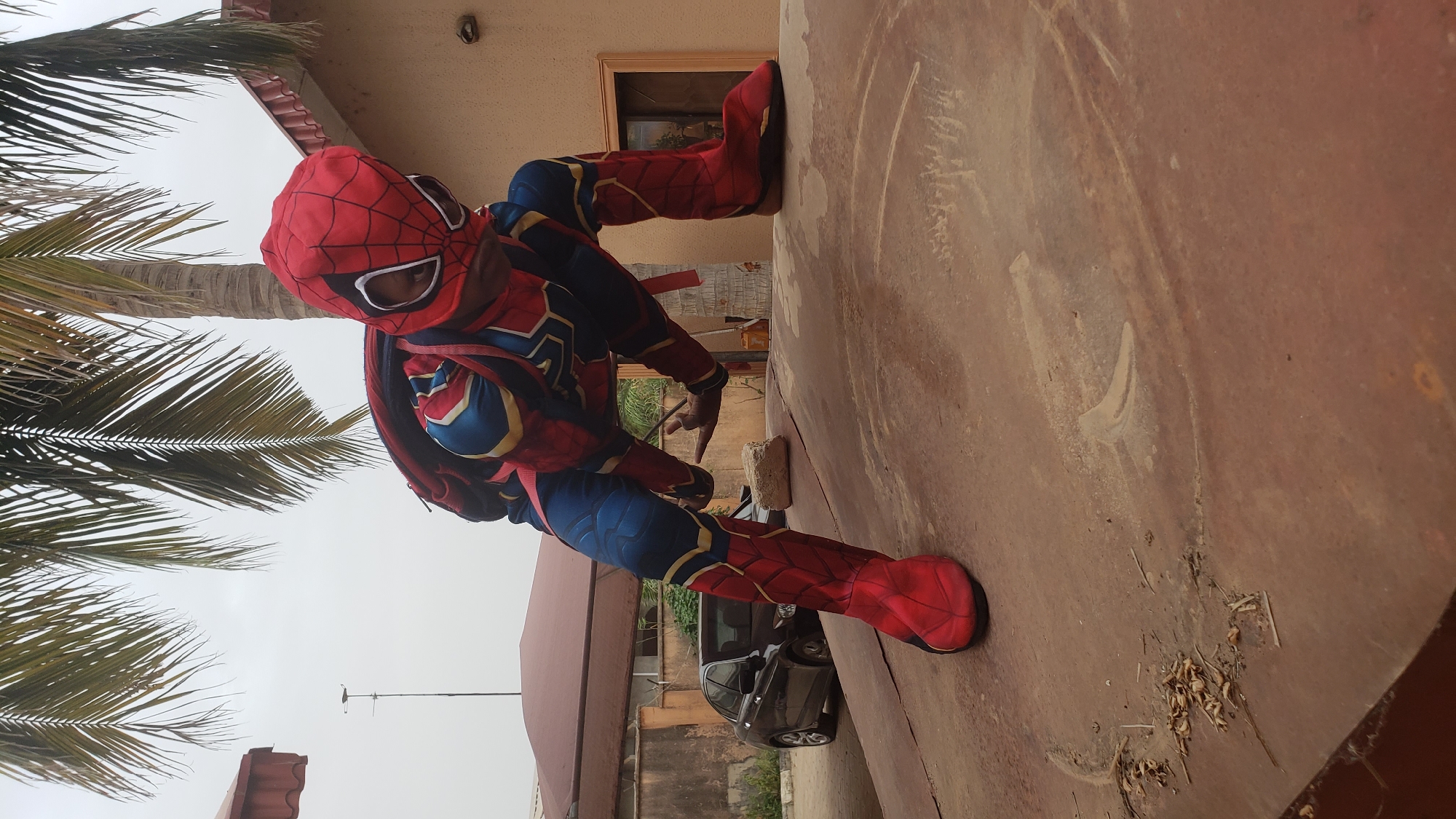 The image shows a person dressed in a Spiderman costume. The person is crouched on a rusty, likely metal surface. A tree is visible to the left of the individual, and a building with a brown door frame is situated behind the person. A grey car is partly visible in the background. Debris, including dry leaves, is scattered on the surface. The photo may have been taken in Sabon Tasha, Nigeria.