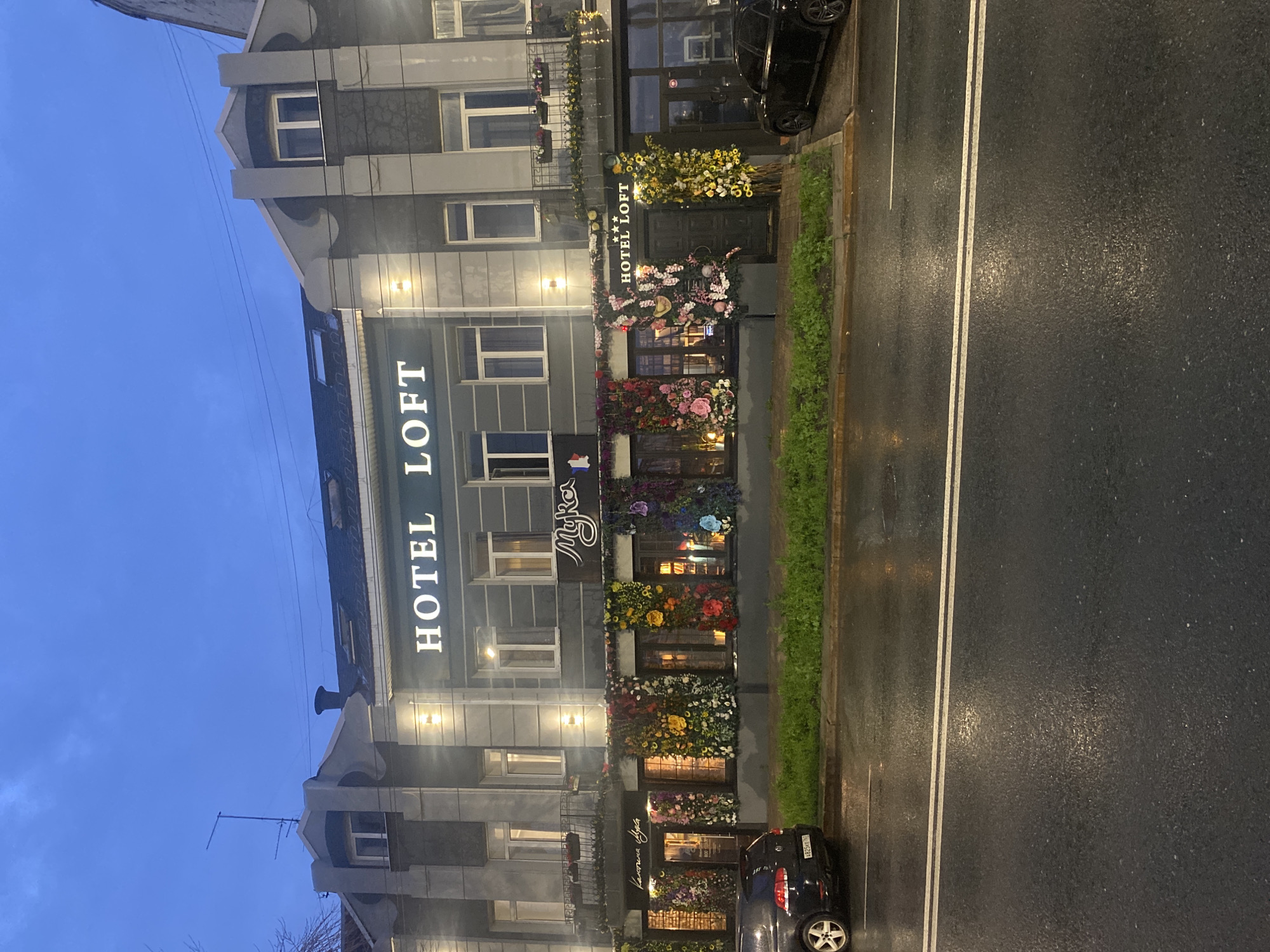 The image shows the facade of the Hotel Loft in Novokuybyshevsk, Russia, at dusk or early evening. The building is a multi-story structure with a dark gray exterior and accented architectural details. The name "HOTEL LOFT" is prominently displayed in white, illuminated letters on the upper part of the building.
The ground floor of the hotel is heavily decorated with vibrant floral arrangements in various colors, including pink, yellow, red, and purple. These flowers are displayed in large clusters and appear to be artificial. There are multiple entrances and windows along the ground floor, some of which are visible through the floral displays. One entrance has signage that reads "Myka" in a cursive font. Another entrance, to the right, also reads "HOTEL LOFT" above the doorway, with decorative sunflowers flanking it.
In front of the hotel, a wet asphalt road reflects the building's lights, indicating recent rain. A black sedan is parked on the left side of the road, and another black car is partially visible on the right. A grassy median runs along the edge of the road, separating it from the hotel's sidewalk. The sky above is a deep blue, characteristic of twilight. The overall scene suggests a well-decorated and somewhat festive atmosphere for the hotel.