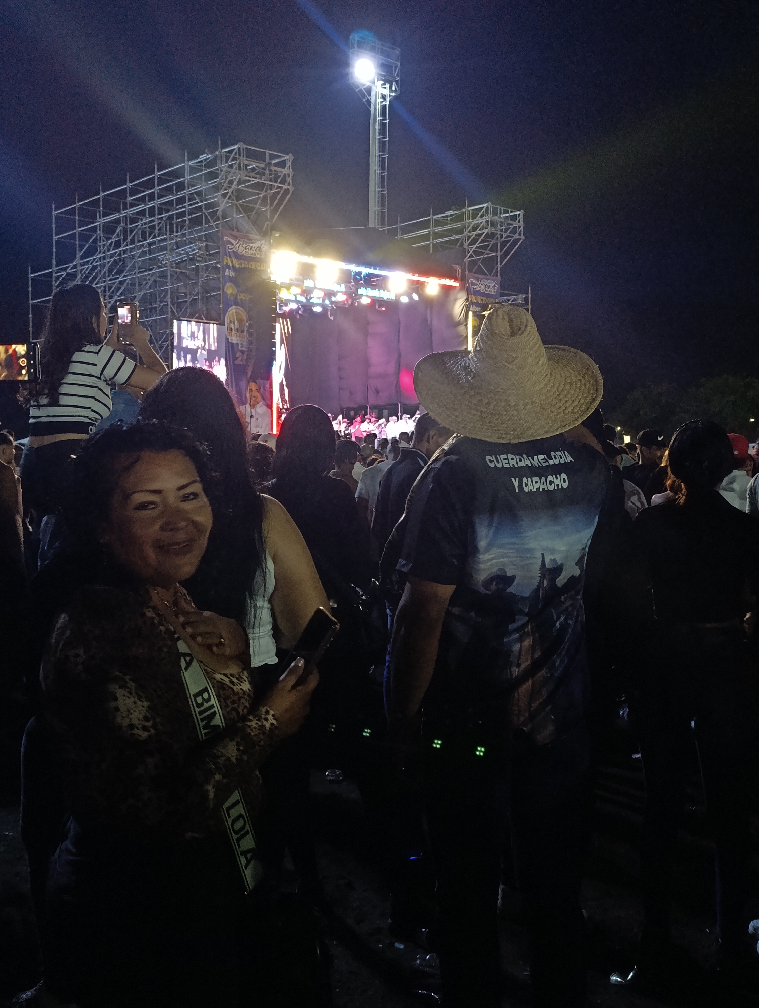 The image captures an outdoor concert or festival at night in Tinaquillo, Venezuela. Bright stage lights and spotlights illuminate the scene, cutting through the dark sky.
In the foreground on the left, a woman wearing a leopard print top and a sash with the partially visible text "BIM LOLA" smiles warmly towards the camera, holding a smartphone. Behind her, another woman with long dark hair is visible, and further back, a woman in a striped shirt records the stage with her phone held high.
In the center-right, the back of a man wearing a wide-brimmed straw hat and a dark t-shirt is visible. His shirt features white text that reads "CUERDAMELODIA Y CAPACHO" above a graphic depicting figures under a sky.
The background is dominated by a large, brightly lit stage structure made of metal scaffolding. Numerous stage lights are active, casting beams into the night. A large screen on the stage displays what appears to be a performer or speaker. A banner hanging near the stage is partially legible, showing "Leyenda" and "PROYECTO CRISTIAN ALBERTO". A large crowd of people, mostly visible as dark silhouettes, fills the area in front of the stage, indicating a well-attended and lively event. The smiling woman in the foreground suggests a festive and enjoyable atmosphere.