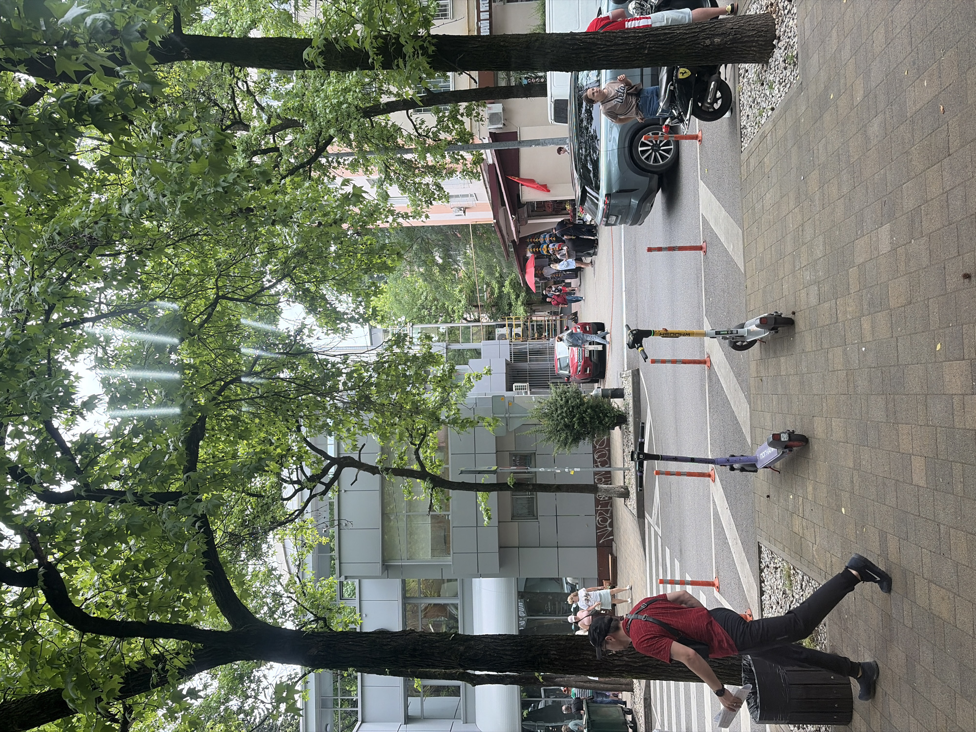 The image depicts a street scene in Sochi, Russia. In the foreground, a man in a red shirt and black pants walks to the left, holding a newspaper and wearing a black bag slung across his chest. To his right, two electric scooters are parked on the sidewalk. The sidewalk is paved with brick-like tiles. Beyond the sidewalk is a street with several parked cars and a motorcycle. In the background, there are buildings, trees, and a red umbrella over a market stall. Several people are visible walking and standing on the sidewalk and in the street. The overall atmosphere is that of a bustling urban environment on a cloudy day.