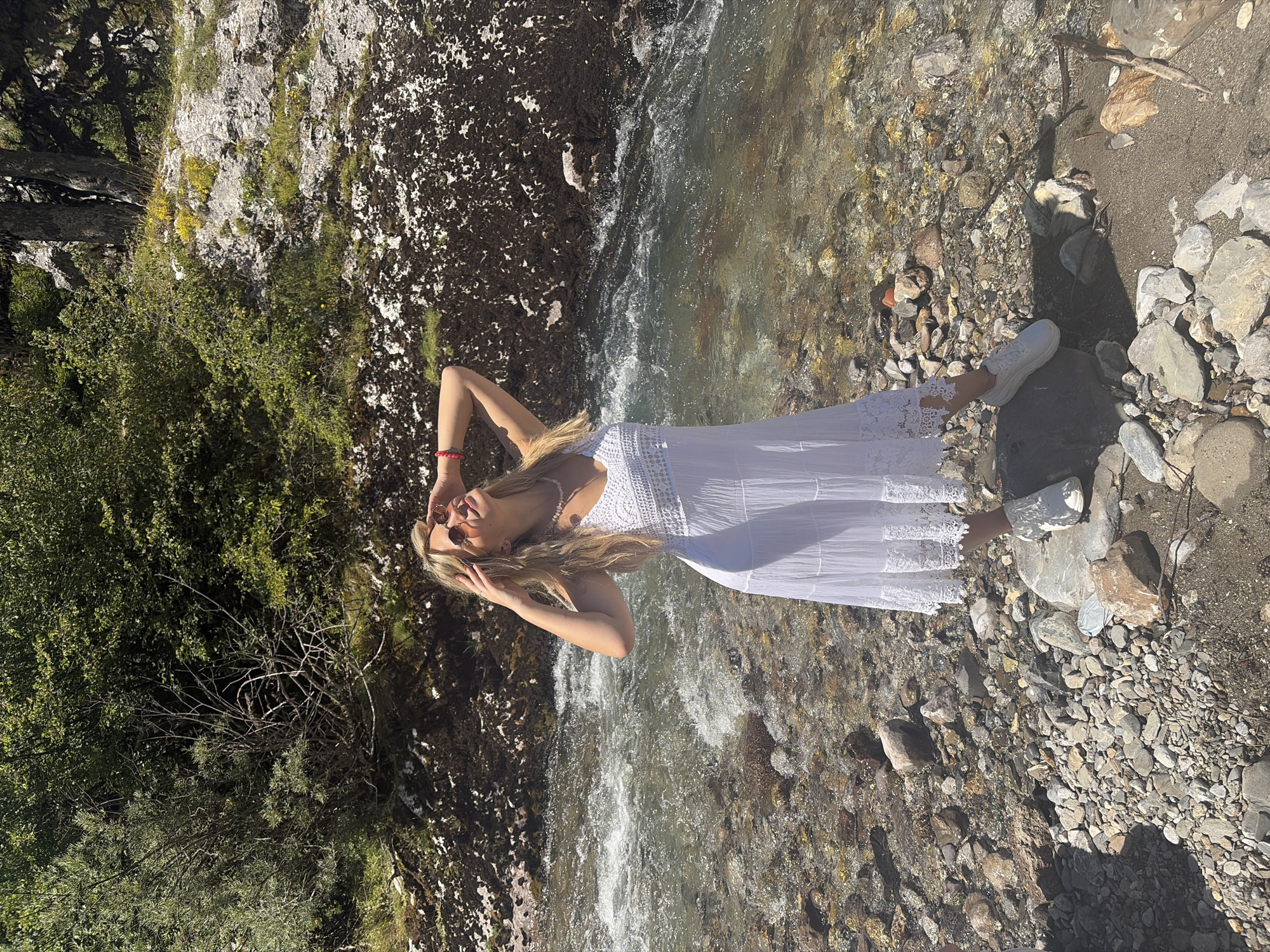 A young woman with blonde hair and sunglasses stands on a rocky riverbank in Vusanje, Montenegro. She is wearing a white lace-trimmed dress and white sneakers. Her hands are raised to her head as she looks up towards the sun. The river behind her flows over a rocky bed, and lush green vegetation covers the hillside. The scene is bathed in bright sunlight, creating a warm and natural atmosphere.
