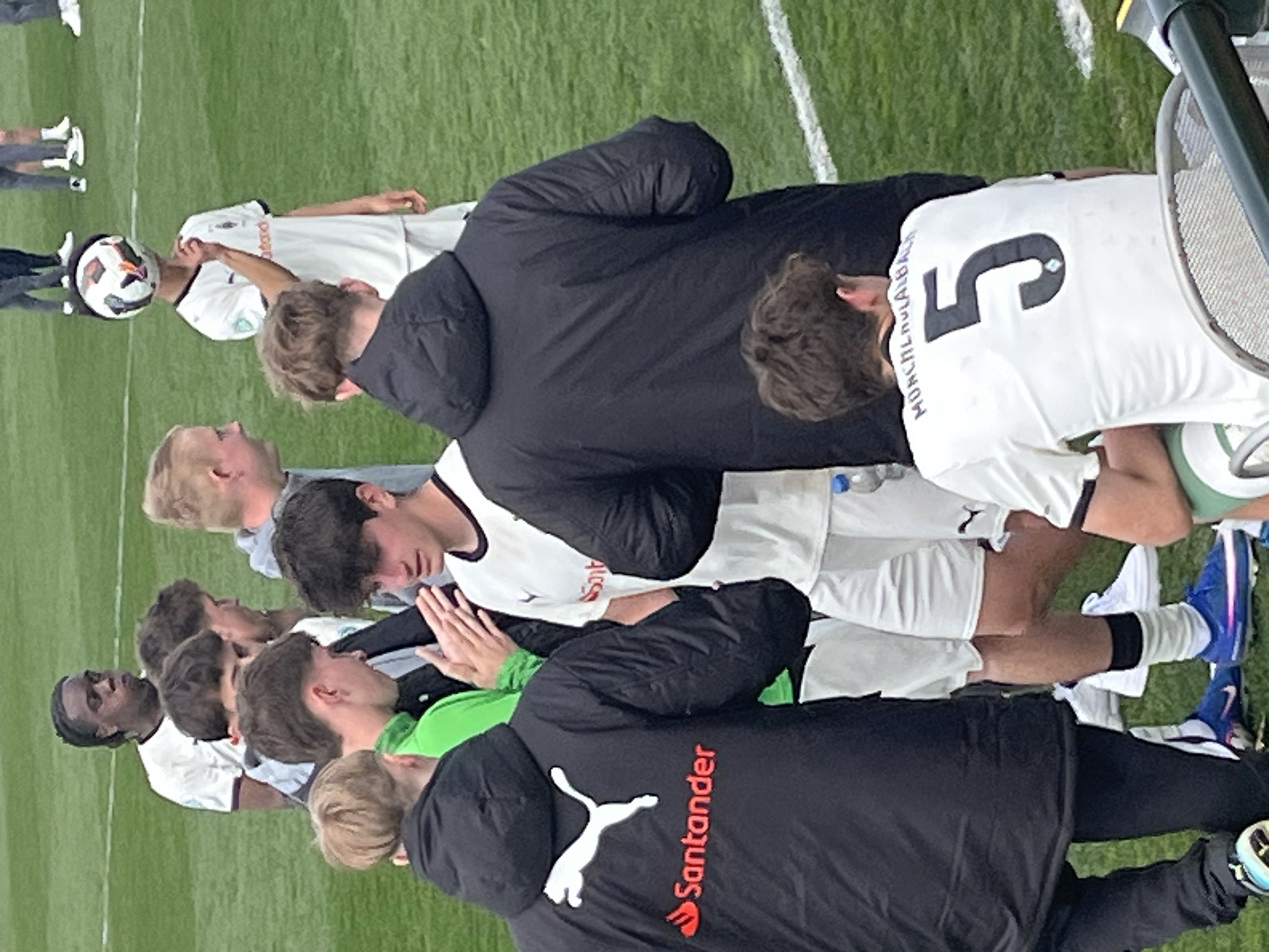 A group of soccer players stand shoulder-to-shoulder in a huddle on a grass field. Some wear white uniforms with black trim, adorned with sponsor logos like "Emirates" and the Puma logo, as well as the number "5" and the name "Monchengladbach" on the back. At least two individuals wear black jackets with the "Santander" logo and the Puma symbol. A soccer ball is visible near the left side of the frame. The setting is likely a soccer field in Mönchengladbach, Germany, due to the team name printed on the back of the jersey. The players appear to be in a moment of team bonding or strategizing.