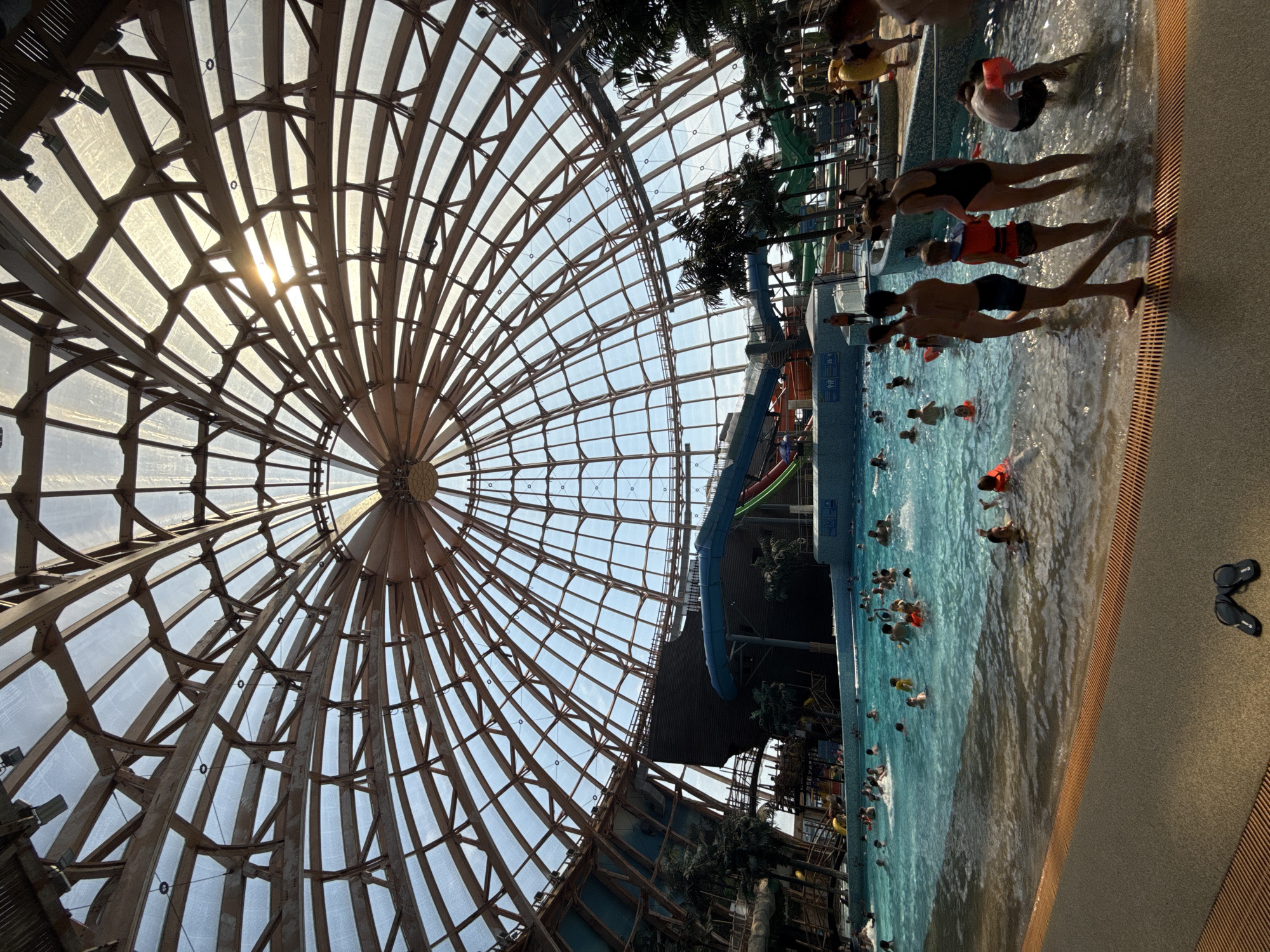 The image depicts a crowded indoor water park under a large, domed, wooden-beamed ceiling with translucent panels that allow sunlight to filter through. A wave pool occupies the foreground and middle ground, filled with numerous people, including adults and children. Several individuals are swimming, while others stand in the shallow areas. In the background, a pirate ship-themed structure with waterslides is visible. Palm trees and other tropical vegetation are interspersed throughout the park, enhancing the atmosphere. The scene suggests a lively and recreational environment. The location is the "Piterland" aquatic center in Saint Petersburg, Russia.