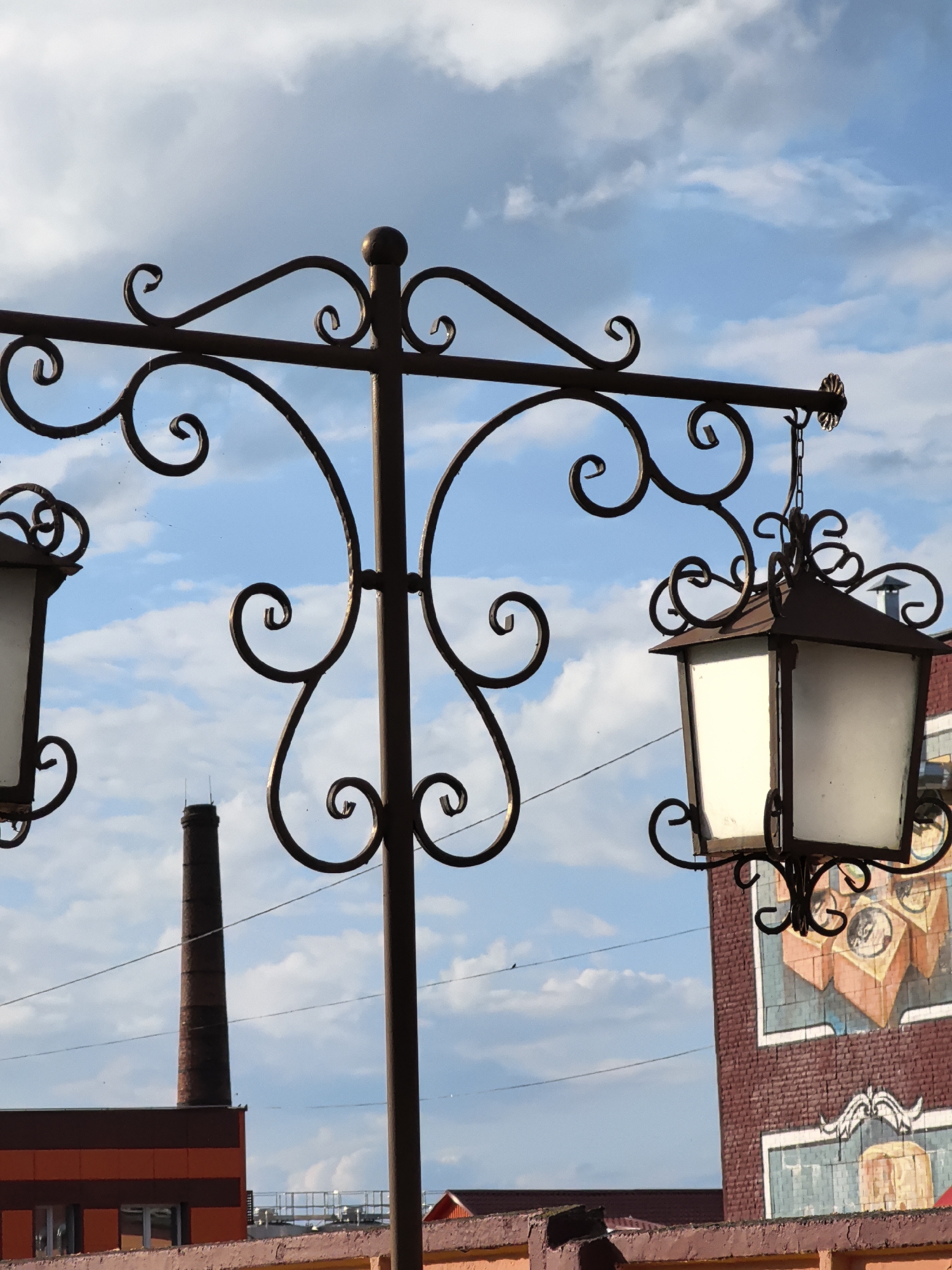 This is a medium-shot, eye-level photograph of an ornate streetlamp against a sky with scattered clouds. The streetlamp has a dark, wrought-iron design with decorative scrollwork. It holds at least two lamps, though only one is fully visible on the right side of the frame. The visible lamp has a square, opaque white pane and a dark, pitched roof.
In the background, a tall, red brick smokestack rises above a building with orange and dark red horizontal siding. Further to the right, part of a brick building with a mural is visible. The mural appears to depict abstract geometric shapes and circular motifs. The overall scene suggests an outdoor urban or industrial setting, possibly with historical or artistic elements. The sky is a clear blue with soft, white clouds, indicating a daytime setting, likely morning or afternoon, with fair weather. There is no visible text in the image. The photograph conveys a sense of old-world charm due to the streetlamp's design, contrasting with the more modern or industrial elements in the background.