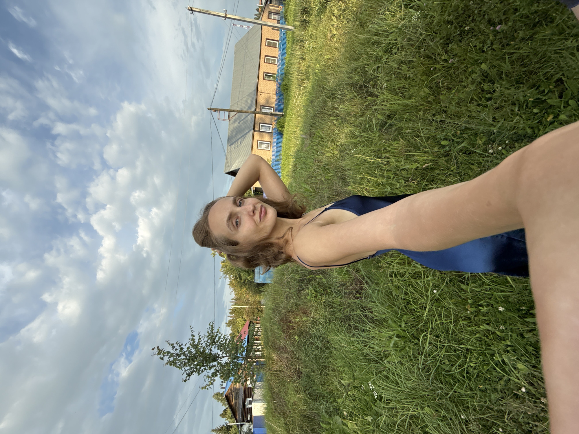 A young Caucasian woman with blonde hair tied up in a bun and wearing a dark blue satin dress is taking a selfie outdoors in a rural village. She is standing in a field of tall green grass. Behind her, in the background, are several traditional Russian wooden houses and a larger brick house with a blue fence. The sky is cloudy with patches of blue. The scene appears to be in the village of Sharkan, Russia, given the architectural style and natural surroundings.