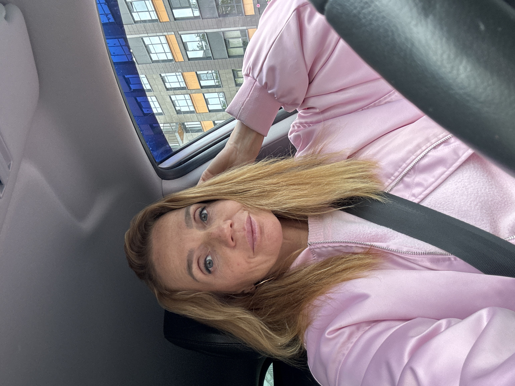 A woman with blonde hair sits in a car wearing a pink jacket and a seatbelt. The car appears to be in the city, with a building visible through the window with multiple windows and some orange accents. The location could be Saint Petersburg, Russia.