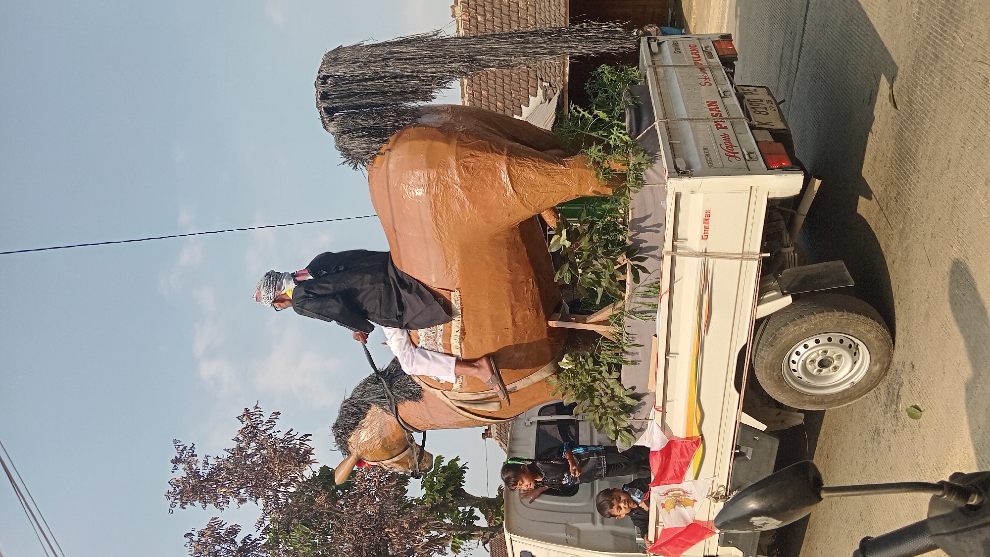In Tinapan, Indonesia, a white pickup truck is the platform for a large, brown, horse-shaped effigy, adorned with a dark, shaggy mane. A person dressed in traditional black clothing and a striped head covering sits atop the effigy, appearing to hold reins. Two young children are visible in the truck bed, one standing and looking towards the left, the other seated and looking downwards. The truck is decorated with red and white flags. The background features trees and a cluster of houses with tiled roofs, suggesting a rural or semi-urban environment. The sky is a clear blue with scattered clouds. The truck's license plate is visible, reading "K 8190 PE". The scene appears to be part of a local procession or festival.