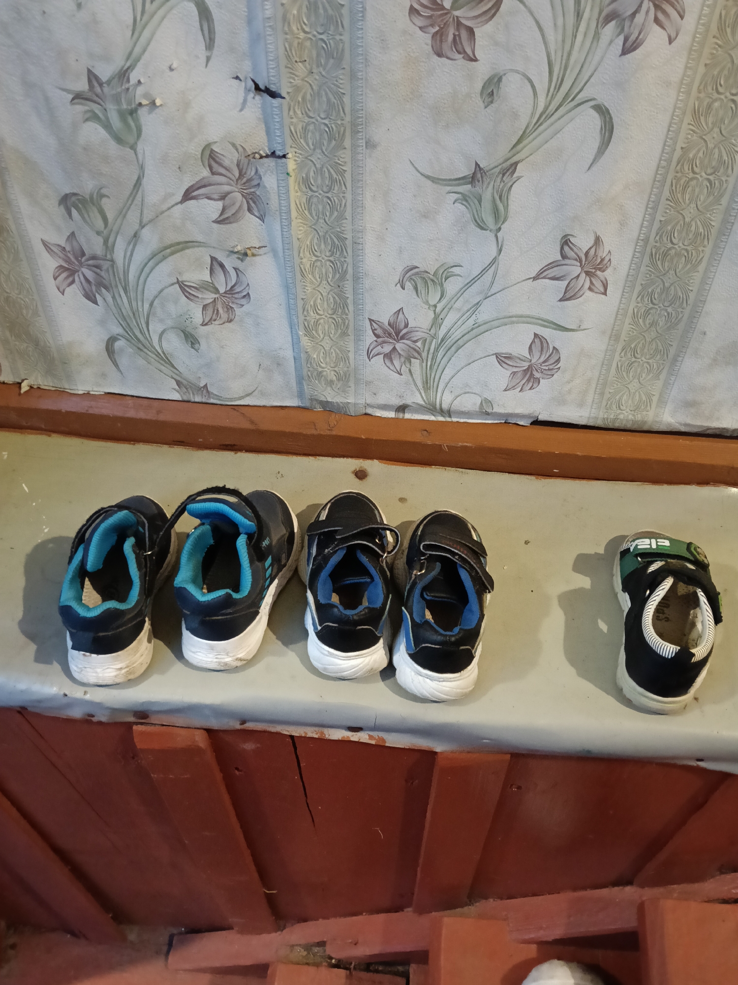 The image displays a line of four pairs of children's shoes resting on a light-colored shelf or ledge. Three of the pairs are dark blue or black sneakers with white soles, featuring light blue interiors and velcro straps. They appear to be well-worn. The fourth pair, on the far right, is noticeably smaller, also dark blue/black with a white sole, and has a distinctive green velcro strap. On this green strap, some white text is partially visible, possibly "213" or part of a brand name.
Below the shelf, a rustic wooden structure, painted a reddish-brown, is visible, suggesting a base or lower part of a wall. Above the shelf, the wall is covered in wallpaper featuring a floral pattern with lilies or similar flowers in muted purple/pink and green tones on a light background. The wallpaper also includes a vertical decorative border pattern. The wallpaper shows signs of age and wear, with some tears and discolored spots.
The scene appears to be an interior or semi-enclosed space, possibly a hallway, entryway, or porch of a home in Красный Маяк, Russia. No people are present, and no specific activity is taking place other than the shoes being stored. The lighting is even, suggesting indirect light. The varying sizes of the shoes indicate multiple children or different growth stages.