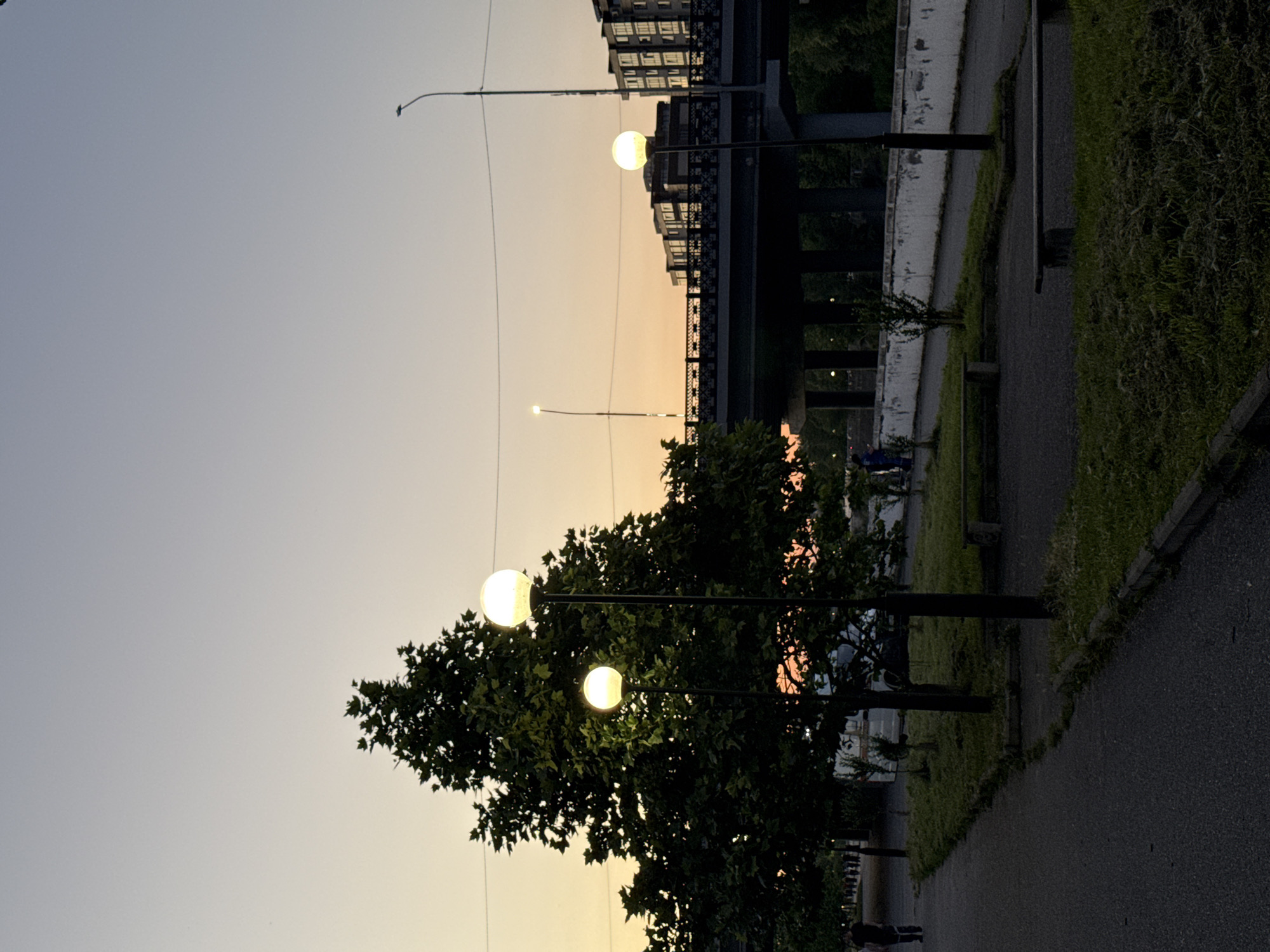 The image captures an outdoor scene at dusk, possibly in Kamysh-Gadzhiaul, Russia. The sky displays a gradient of light blues and pale yellows, suggesting the sun has recently set. Several streetlights with glowing globes are visible, some illuminating nearby trees and a paved pathway. One streetlight in the foreground is particularly prominent, with a large, illuminated sphere. Beyond the pathway, a line of grassy embankment leads to a building with multiple windows, and a dark structure resembling a bridge or overpass stretches across the middle ground. The overall atmosphere is tranquil, with the warm glow of the streetlights contrasting with the deepening twilight. The presence of a pathway and benches hints at a public park or promenade. A few indistinct figures can be seen on the pathway in the distance, suggesting people are present, perhaps enjoying the evening. The vegetation is lush and green, indicating a temperate climate. There is no visible text in the image.