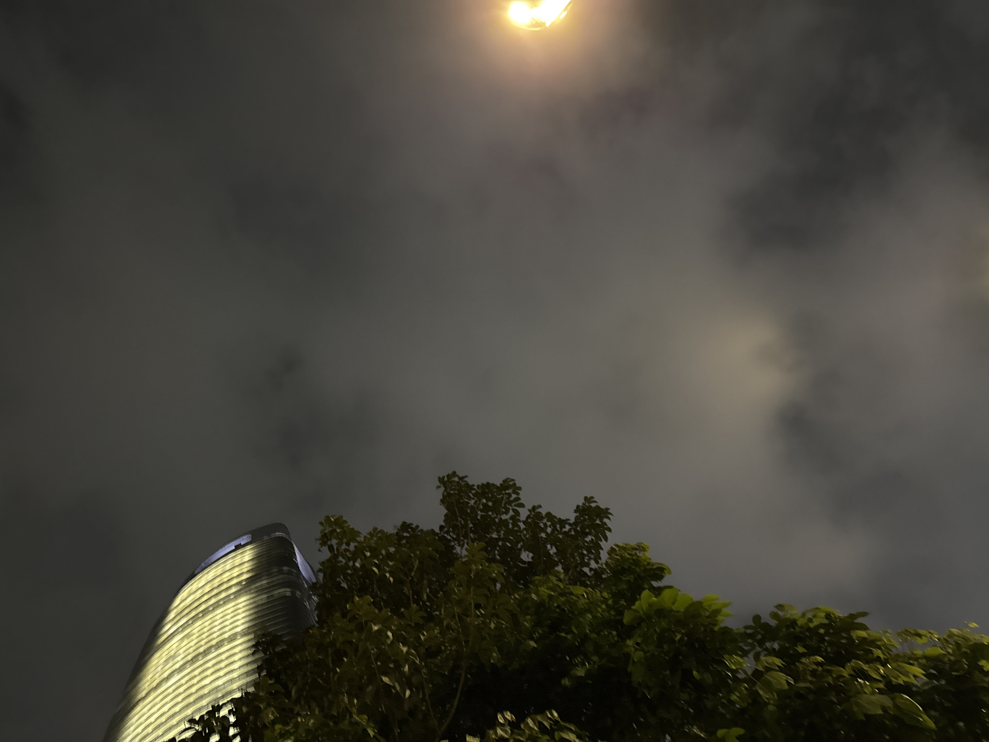 The image captures an outdoor night scene in Haizhu District, China, dominated by a cloudy sky. A bright, yellow light source, likely a streetlamp, is visible at the top of the frame, illuminating the dark clouds with an ethereal glow. Below the sky, a portion of a modern, curved skyscraper is visible on the left, its facade striped with warm, golden lights that reflect off its surface. The building's design appears sleek and contemporary. To the right of the skyscraper, lush green foliage from a tree extends into the frame, its leaves silhouetted against the dimly lit sky. The combination of the illuminated skyscraper and the dark, overcast sky suggests a dramatic and somewhat mysterious urban atmosphere. The scene evokes a sense of a calm, yet perhaps slightly moody, evening in a city. No people or specific activities are depicted, and no text is visible in the image.