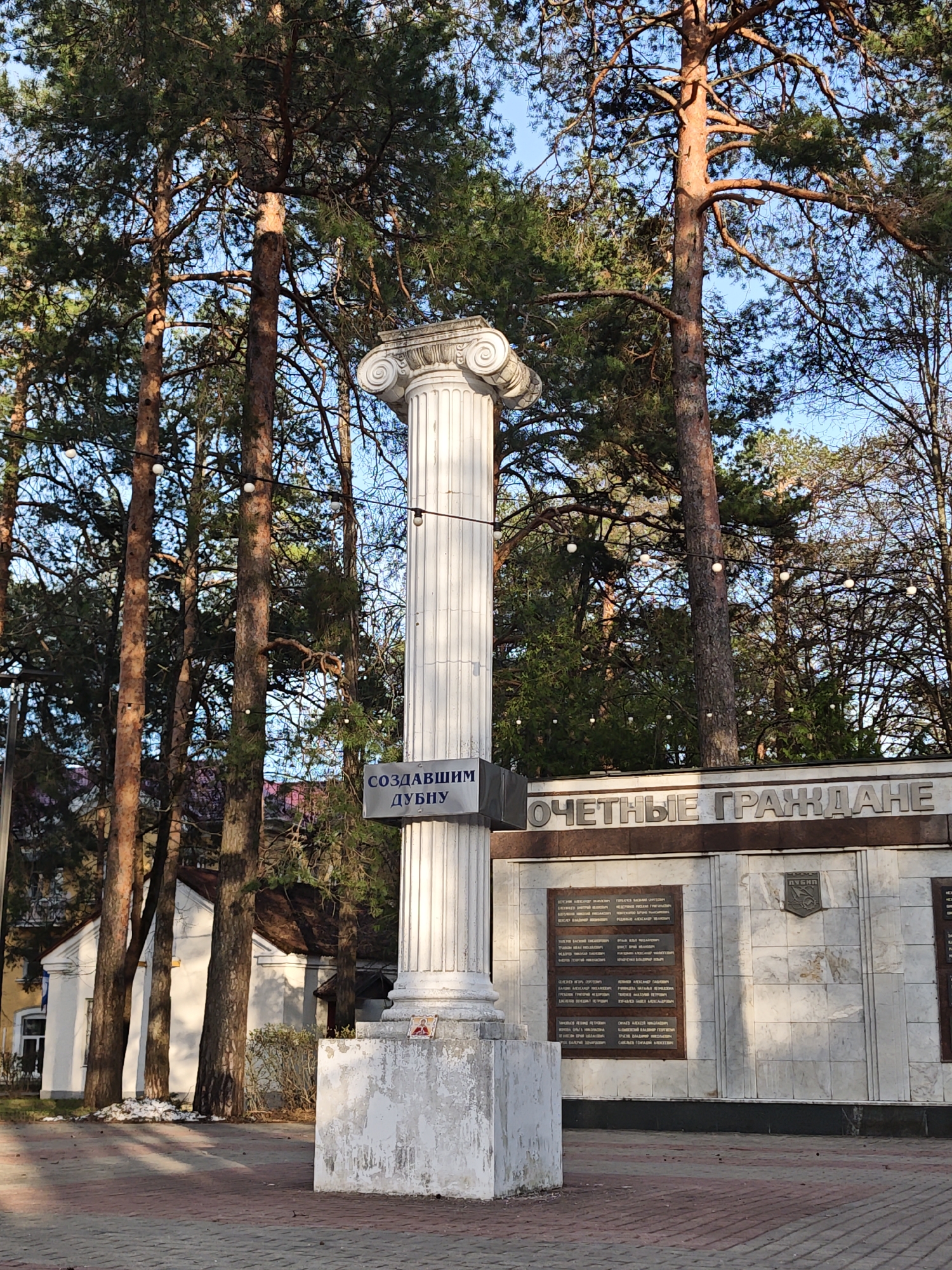 Here's a detailed description of the image:
The image showcases a tall, white, fluted Ionic column standing prominently in what appears to be a public space. A sign attached to the column reads "СОЗДАВШИМ ДУБНУ" in Cyrillic, which translates to "To the creators of Dubna." The column is situated on a weathered, white plinth.
Behind the column is a wall with the inscription "ОЧЕТНЫЕ ГРАЖДАНЕ" (Honored Citizens) with inscribed plaques. The plaques contain lists of names.
The column and memorial wall are set against a backdrop of tall pine trees, which suggests a park-like environment. A string of lights is strung between the trees. The ground is paved with red and gray brick pavers. In the background, a white building with a dark roof and a yellow building can be seen.
The scene is located in Krasnaya Gorka, Russia.