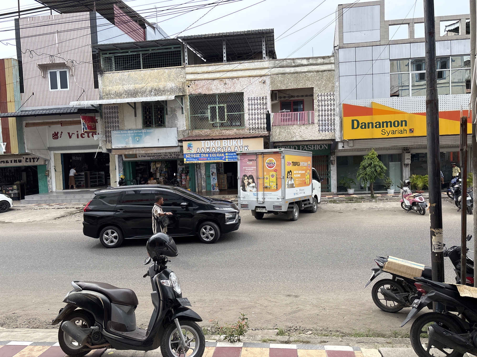 The image depicts a street scene in Lhokseumawe, Indonesia, with several buildings lining the road. In the foreground, a black scooter with a helmet secured to its handlebars is parked on the side of the road. Further down the street, a black SUV is stopped, and a man in a striped shirt is standing beside its open driver's side door. A white truck with advertising graphics is also present on the road.
The buildings appear to be commercial establishments. One building prominently features a large yellow and orange sign for "Danamon Syariah". Another building has signs for "VICTORY" and "TOKO BUKU JAKARTA ATK". There are also signs with text in Indonesian. The buildings have various architectural styles, with some appearing older and others more modern.
The street is paved, and there is a sidewalk with patterned tiles in the foreground. Various trees and potted plants are visible along the sidewalk and in front of the buildings. The sky is overcast. Several other motorcycles are parked in the background.