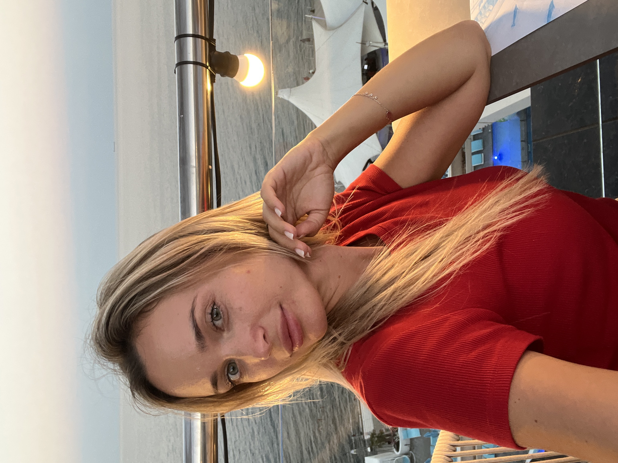 A young blonde woman with blue eyes and fair skin is captured in a close-up, waist-up selfie, posing outdoors with a serene ocean backdrop. She is wearing a vibrant red, ribbed t-shirt. Her hair, a cascade of blonde, falls over her left shoulder, with some strands framing her face. Her right arm is bent, with her hand resting delicately on her chin, and a thin bracelet adorns her wrist. The woman's gaze is directed at the camera, conveying a relaxed and pleasant demeanor.
Behind her, the vast expanse of the sea stretches to the horizon, merging with a pale sky painted in hues of pink and blue, suggesting either sunrise or sunset. A sleek metal railing runs horizontally across the frame, separating the woman from the view. A single, illuminated bulb hangs from a wire above the railing, casting a warm glow. In the background, partially obscured by the railing and the woman's arm, are glimpses of white tent-like structures, hinting at a beachside resort or event space. The presence of a swimming pool with illuminated blue water is also visible below. The overall scene appears to be a tranquil setting by the sea, likely within Odesa, Ukraine, given the context provided.