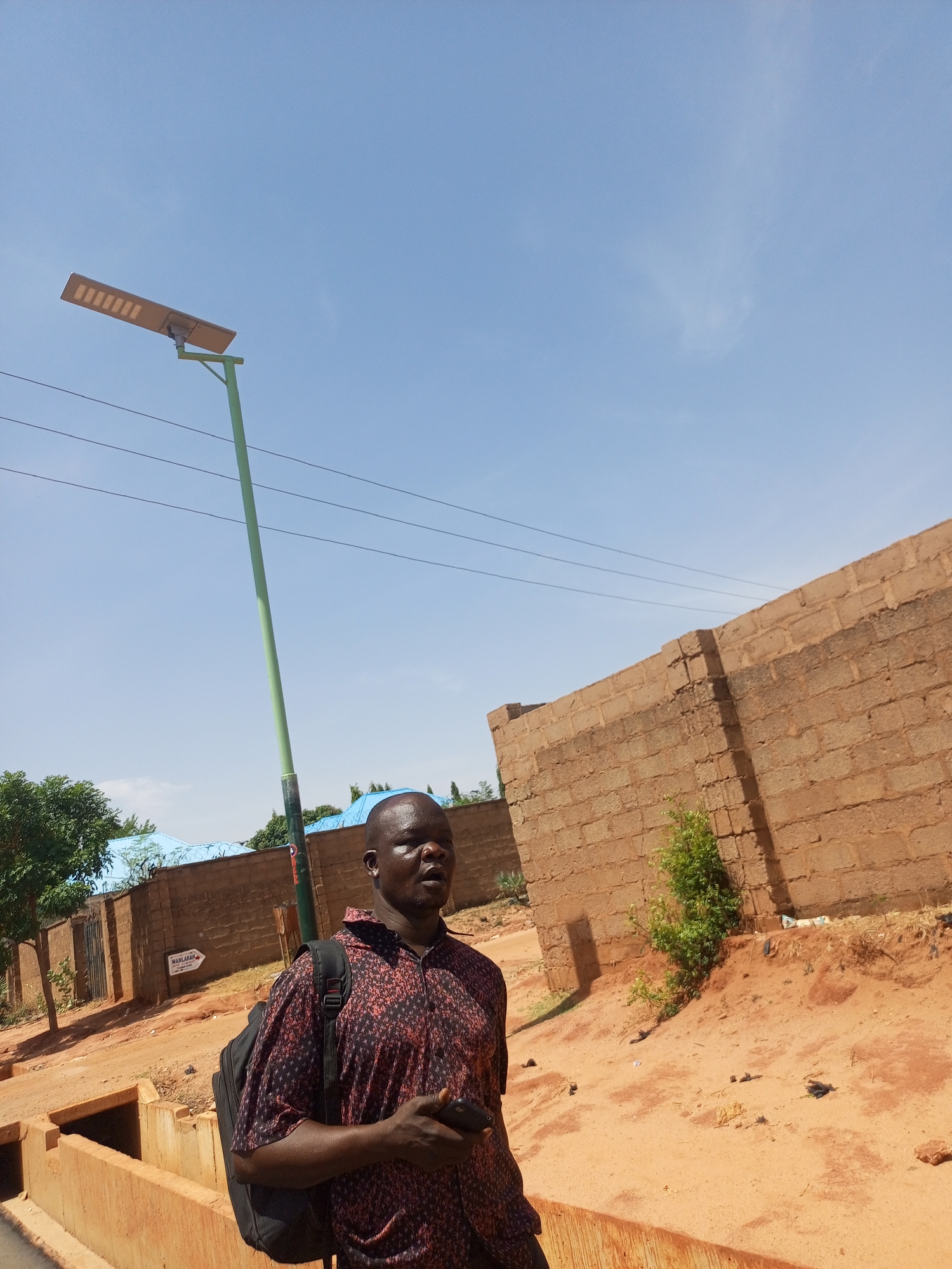 An outdoor, eye-level shot on a bright, sunny day features a man standing on a dirt path next to a concrete drainage ditch. The man, with dark skin and short hair, wears a patterned short-sleeved shirt (dark red/black) and a black backpack slung over his left shoulder. He holds a dark-colored smartphone in his right hand and looks slightly to his right with a neutral expression.
The setting appears to be a residential or semi-urban area. The ground is reddish-brown earth/sand. To the man's right, a tall wall made of weathered, light brown concrete blocks runs along the path, with a small green bush growing at its base. To his left, beyond the ditch, are green trees, a fence with barbed wire, and some buildings with blue corrugated metal roofs.
A tall, light green solar street light with a rectangular fixture is prominently positioned in the left mid-ground, extending towards the clear, bright blue sky which dominates the upper portion of the image. Overhead power lines are visible crossing the sky behind the streetlight. In the mid-ground, on the left side of the path, a white rectangular sign with an arrow pointing left is attached to a fence or structure. The sign clearly displays the text "WAMLARAH", with some smaller, less legible text below it. The shadows are relatively short, indicating midday or early afternoon.