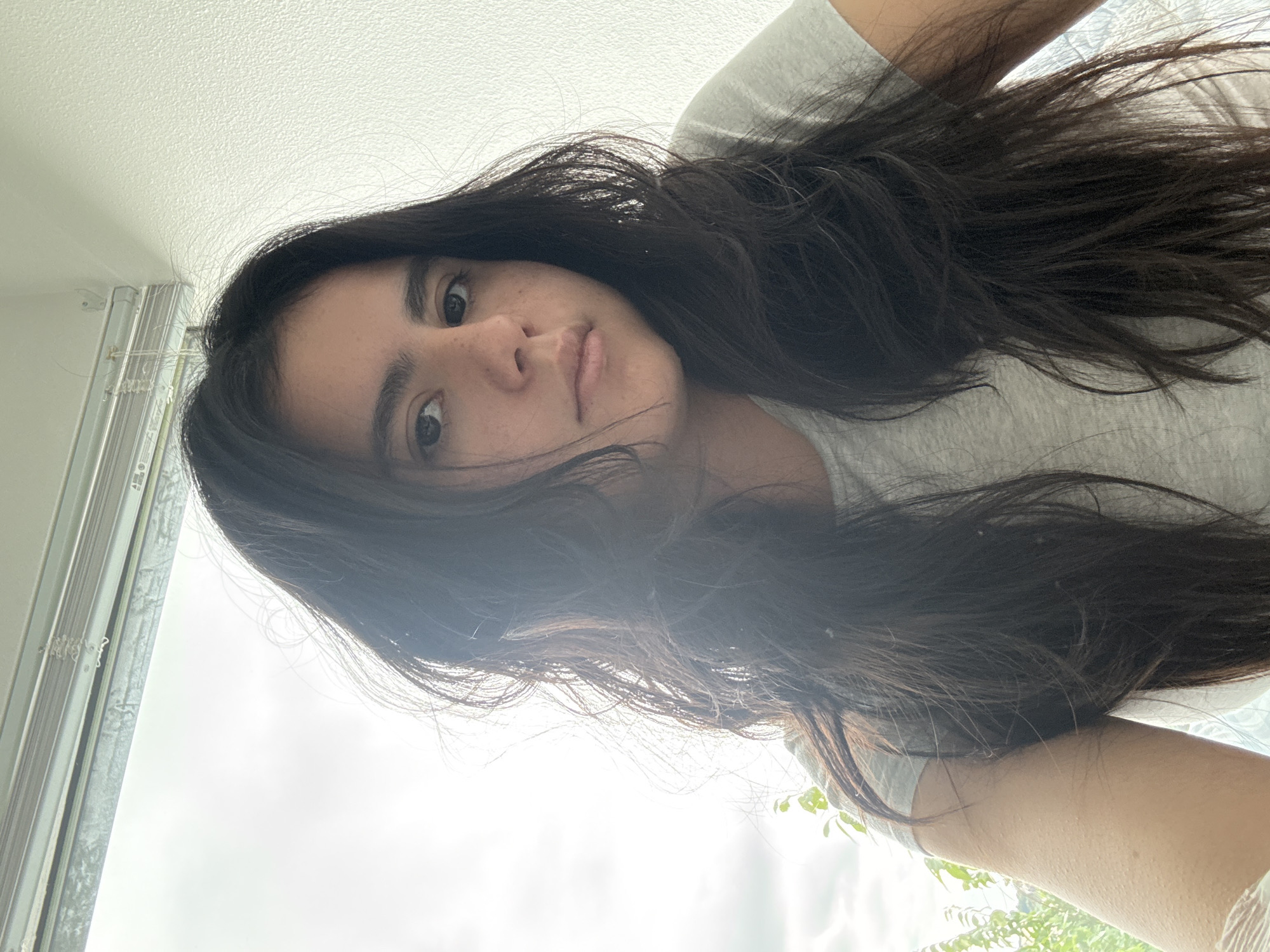 The image features a young woman in a close-up portrait, oriented such that her head is to the left and her body extends towards the right. She has long, dark, wavy hair that frames her face and falls across her shoulders and chest. She is wearing a light-colored t-shirt. Her face is visible, looking directly at the camera with a neutral, calm expression. Her dark eyes, natural eyebrows, and freckles across her nose and cheeks are clearly visible.
The background is a very bright, predominantly white area, likely an overcast sky or bright ceiling, indicating daytime. On the far left of the image, vertical window blinds or a window frame are visible. Faint green foliage can be seen at the bottom right, suggesting an outdoor view beyond a window. The camera appears to be positioned below the woman, looking upwards. This casual photo was taken in Austin, United States of America. No visible text is present.