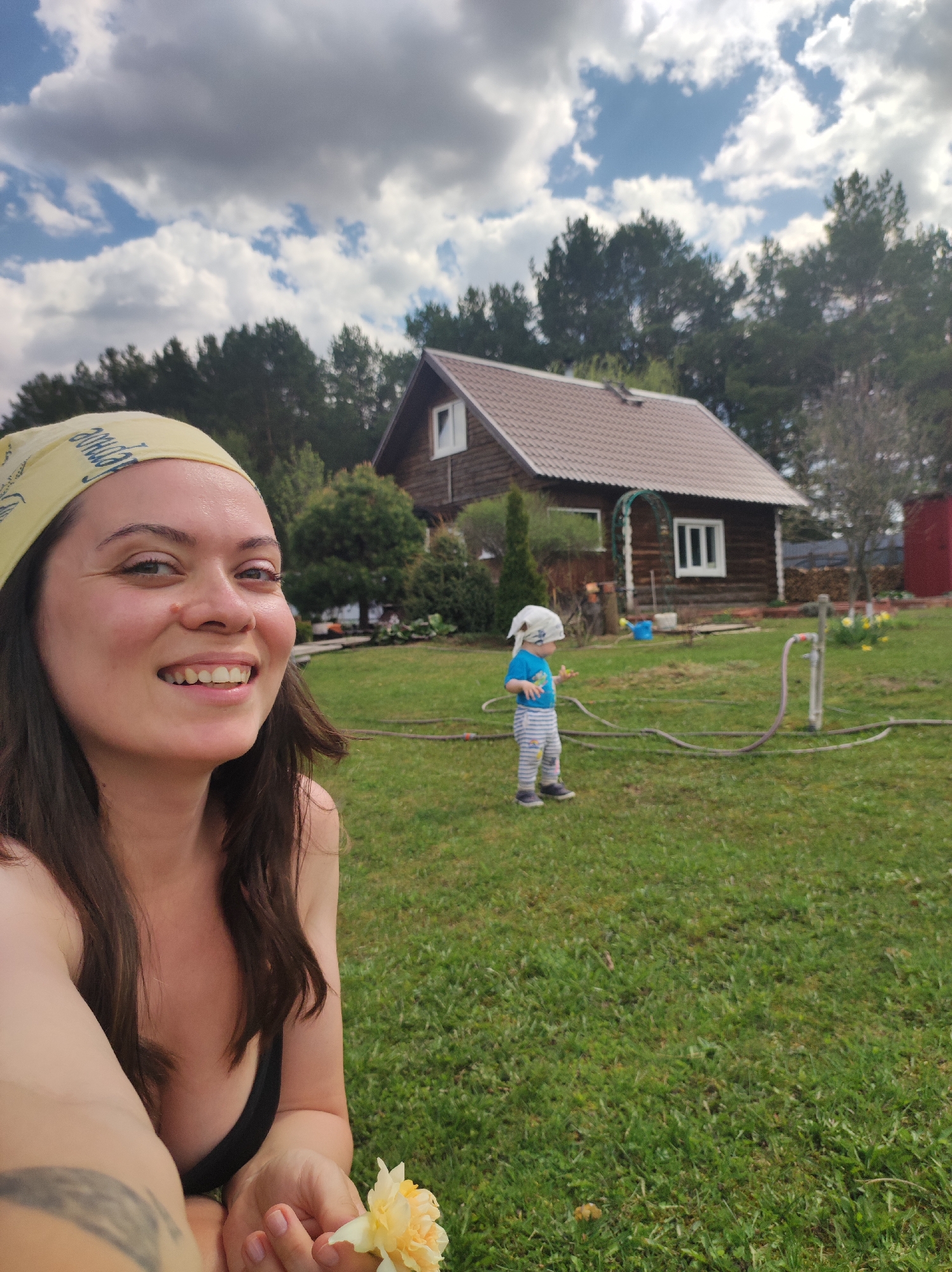 A selfie taken outdoors shows a woman with long brown hair wearing a yellow headscarf, smiling. She is holding a small, yellow flower. In the background, a young child wearing a light-colored head covering, a blue shirt, and striped pants stands on a green lawn. A brown, two-story wooden house with a tiled roof is visible behind the child. Trees surround the house, and there is a partially visible red structure to the right of the house. A garden hose lies on the lawn. The sky is partly cloudy. The location is Фоки, Russia.