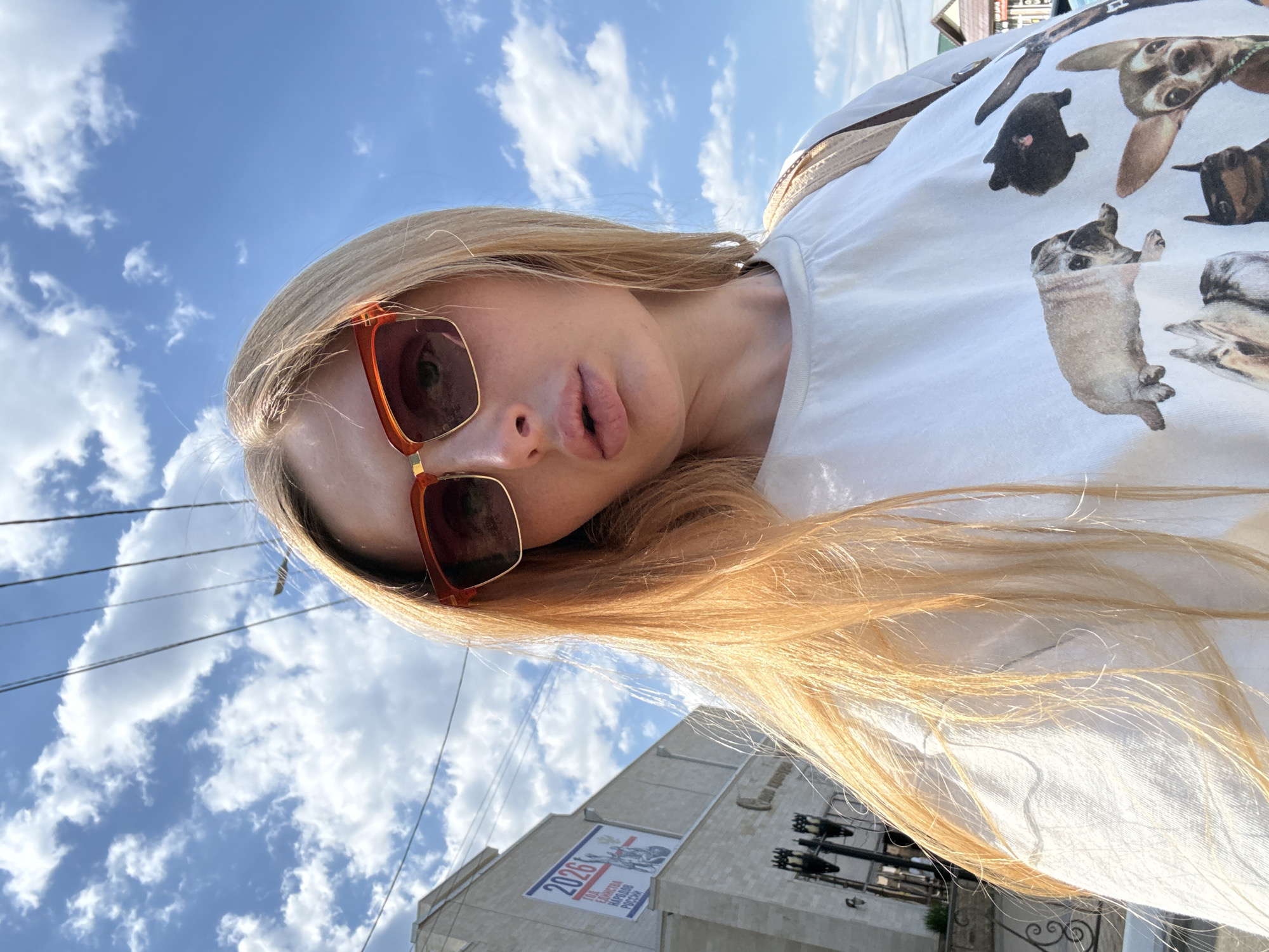 The image shows a close-up, low-angle selfie of a young woman with long blonde hair and sunglasses with orange frames. She is wearing a white t-shirt with a pattern of pug dogs. Her mouth is slightly open, and she is looking towards the camera. The background is a bright blue sky with scattered white clouds. Power lines are visible in the upper left corner. In the lower part of the image, a building with a banner is visible. The banner has text that appears to be in Russian and includes the number "2026". The time of day appears to be daytime, likely sunny, given the bright sky and lighting. The overall mood seems casual and outdoorsy.