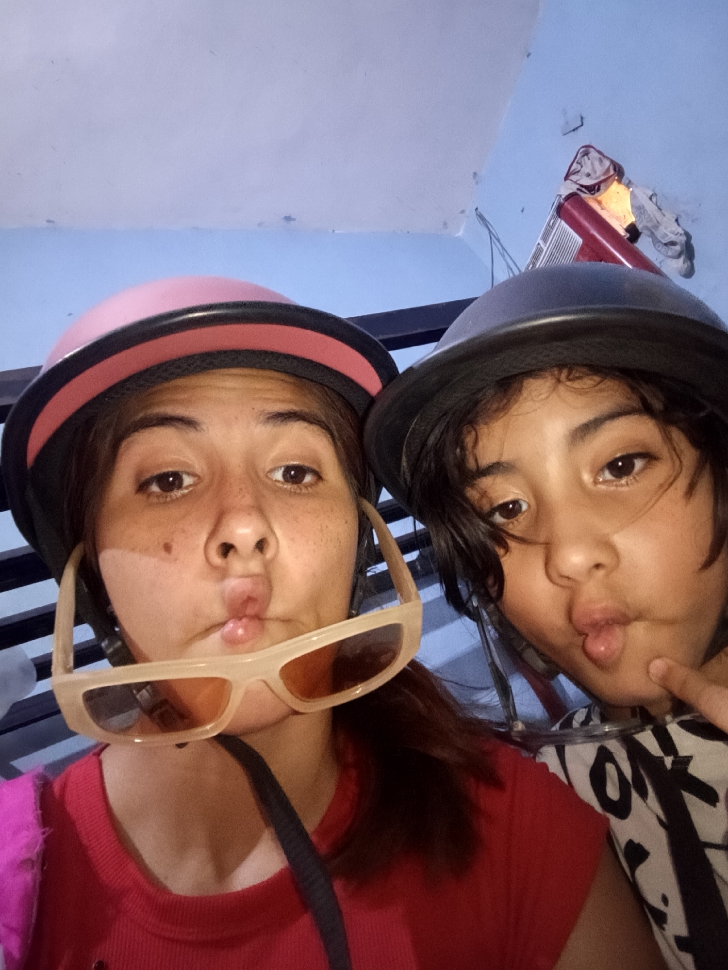Two individuals, likely a mother and child, are positioned closely together, possibly taking a selfie. Both are wearing helmets, the woman's helmet is pink, with sunglasses hanging near her mouth. Both individuals are pouting their lips in a playful manner. The environment appears to be indoors, with a partial view of a wall and a red ladder in the background. There is no clear indication of this being in Juan Griego, Venezuela.