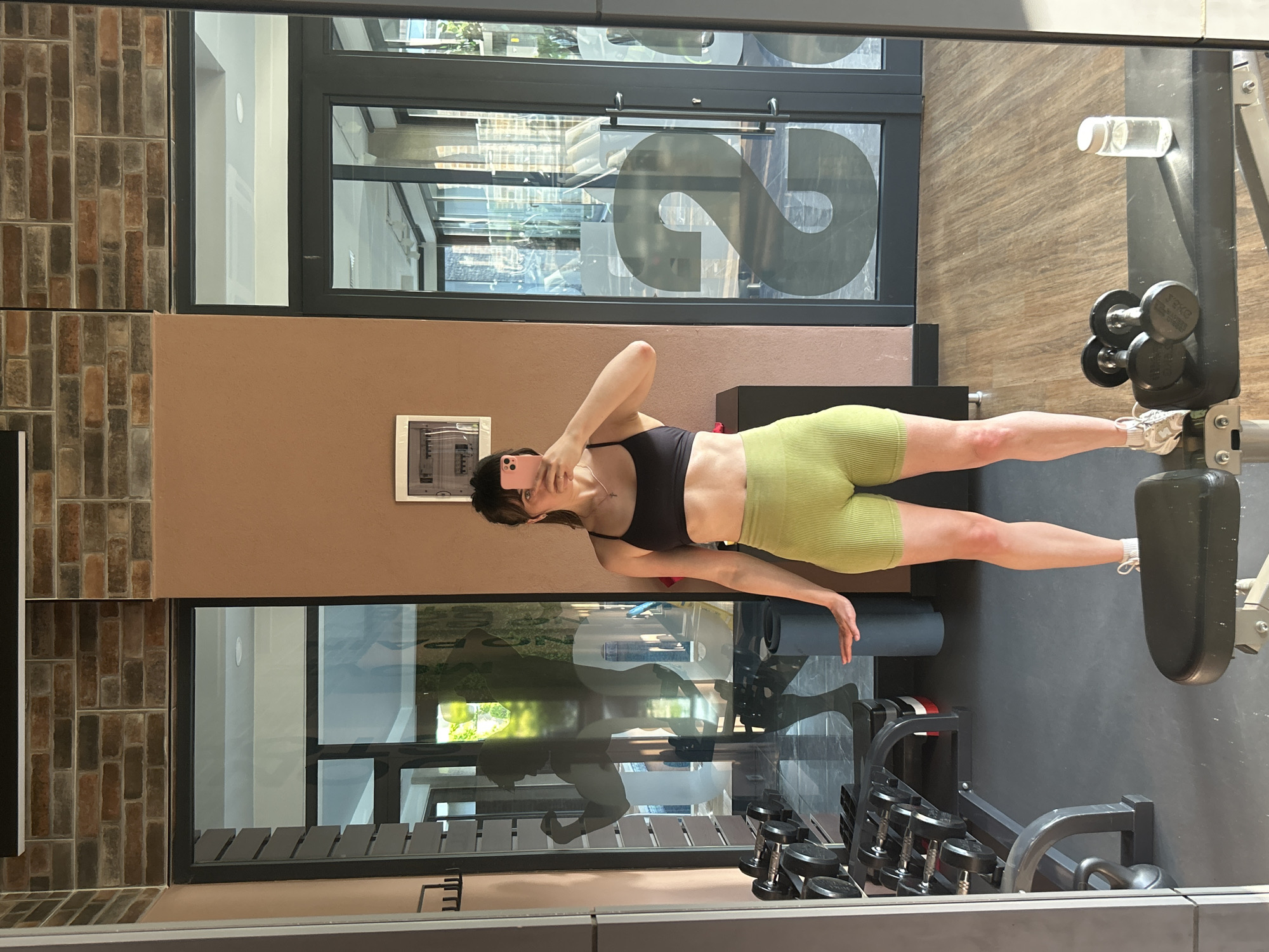 A woman is taking a selfie in a gym setting, presumably in the city of Alanya, Turkey. She is wearing a black sports bra and light green ribbed shorts. The gym has brick and tan colored walls and large windows with black frames. Weights and workout equipment are visible.