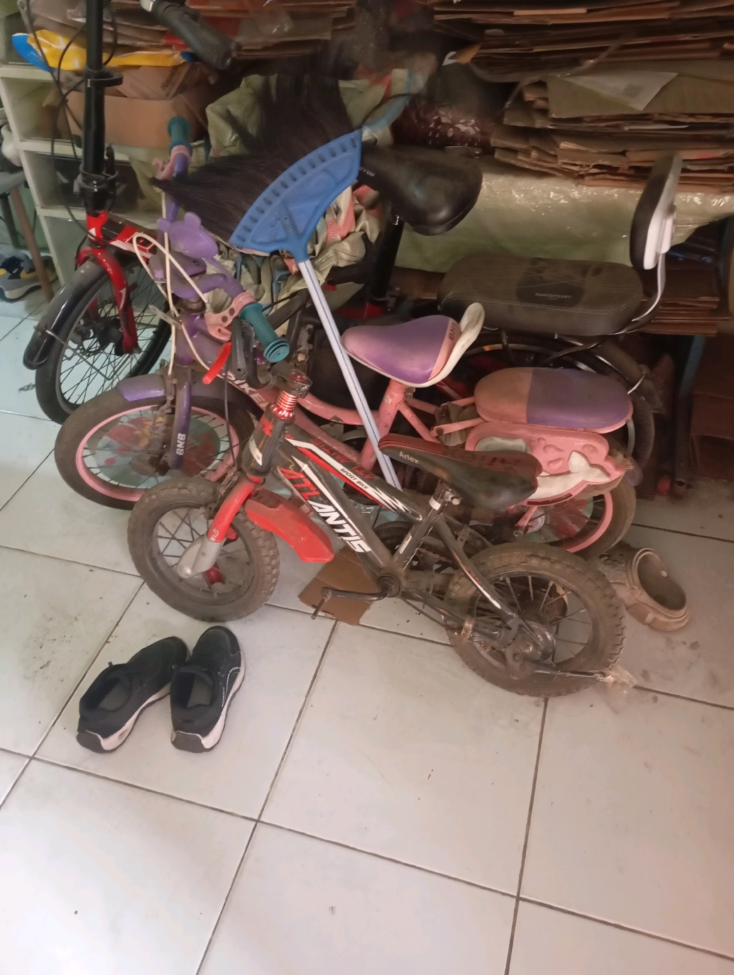 This image depicts a cluttered indoor storage area in Babelan, Indonesia, likely a garage or shed. The primary subjects are two children's bicycles, a smaller red one with a black and red frame, and a larger pink one with purple accents. The red bicycle is positioned in the foreground, facing left, with its front wheel slightly angled. The pink bicycle is behind it and to the left, with its front wheel turned sharply to the right. Both bicycles appear to be standing upright without support.
Beside the bicycles, on the tiled floor, are a pair of black athletic shoes, with the left shoe placed slightly in front of the right. A blue-handled broom with a blue plastic rake head is leaning against the bicycles, partially obscuring the handlebars of the pink bicycle.
The background is filled with stacks of cardboard boxes, some appearing to be flattened and others intact. Various bags and miscellaneous items are also visible, contributing to the sense of disarray. A white shelf unit is partially visible on the far left side of the image. The lighting is dim and appears to be artificial, casting soft shadows on the tiled floor. There are no people visible in the image. The scene suggests a storage space for sporting equipment and household items.