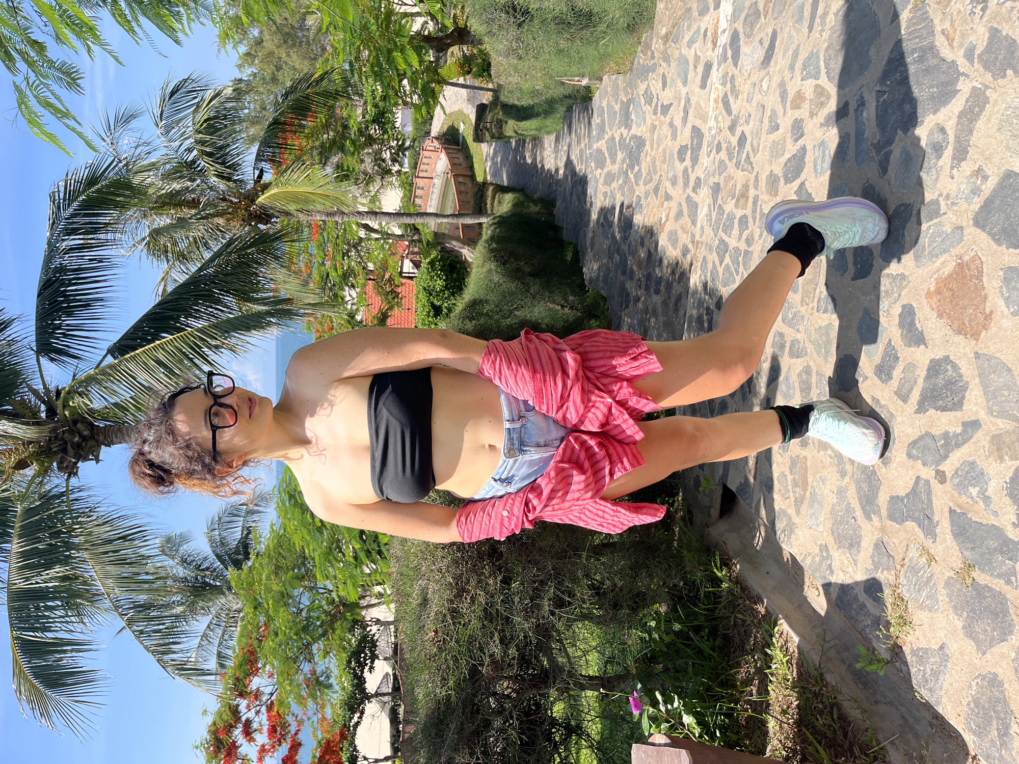 A woman with curly brown hair styled in a messy bun, wearing black-rimmed glasses, stands on a stone staircase in Phu Thuy Ward, Vietnam. She is dressed in a black strapless top, denim shorts, and a pink striped unbuttoned shirt tied at her waist. She has black socks and light blue and purple athletic shoes on her feet. The background features lush green vegetation, palm trees, and a glimpse of the ocean under a clear blue sky. A stone wall and pathway are visible further down the stairs. The scene is brightly lit by sunlight, casting shadows on the stone steps.