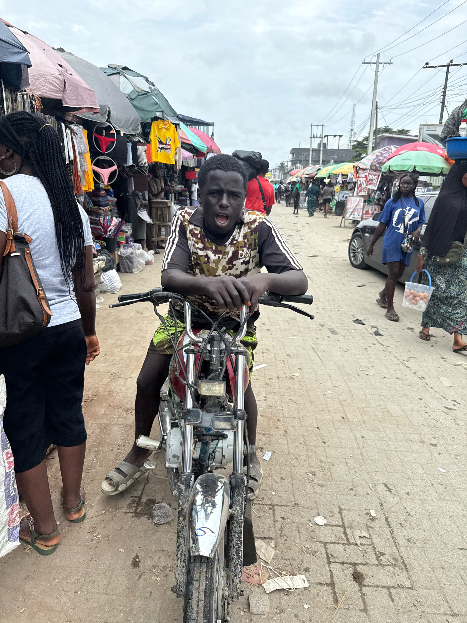 A young Black male, appearing to be in his teens, sits on a dirt-covered red motorcycle in a bustling outdoor market. He is wearing a camouflage-patterned t-shirt with black stripes on the sleeves and green shorts. His mouth is open, and he is looking directly at the camera. The motorcycle has a damaged headlight.
To the left of the young man, a Black female with long braided hair and wearing a grey t-shirt and black shorts is partially visible, facing away from the camera. She is carrying a brown bag over her shoulder.
The background depicts a crowded market scene. Stalls with colorful awnings display various goods, including clothing items like yellow t-shirts and undergarments. Several other people are visible in the distance, some walking and some tending to stalls. A silver car is parked on the right side of the frame, and overhead power lines crisscross the sky, which is overcast. The ground is a dusty, paved surface with scattered debris. The location is identified as the Ajah market in Lagos, Nigeria.