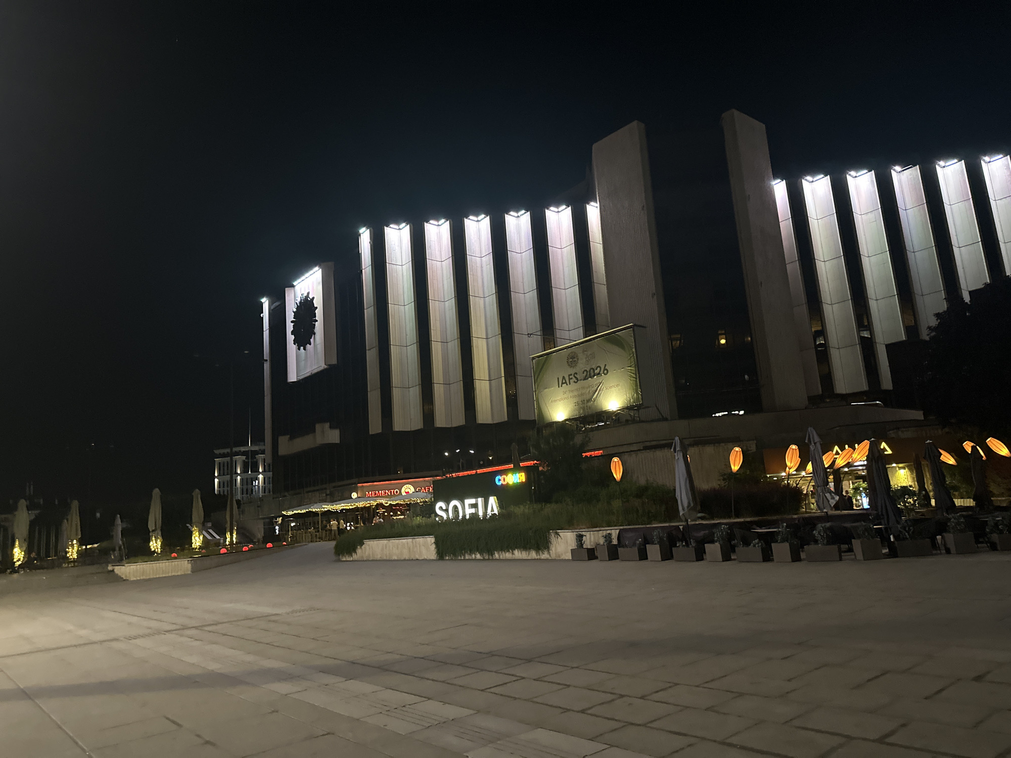 The image shows a night view of the National Palace of Culture (NDK) in Sofia, Bulgaria. The NDK building is illuminated by vertical lights, creating a striped effect on its facade. In the foreground, a paved plaza stretches out, with several closed outdoor seating areas featuring umbrellas and planters. To the left, a sign with the word "SOFIA" in large, illuminated letters is visible, accompanied by the "Memento Cafe" sign. A large banner advertising "IAFS 2026" is also displayed on the building. The sky above is dark.