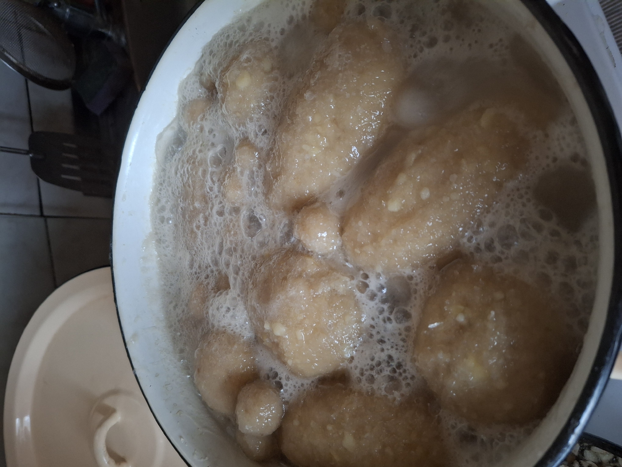 In Traupis, Lithuania, a white enamel pot filled with boiling water contains several light brown, oval-shaped dumplings. The dumplings are partially submerged in the bubbling water, with some pieces of what appear to be finely minced garlic visible on their surfaces. The pot is positioned on a stovetop, with a beige lid of a similar material nearby. In the background, a dark tiled wall is visible, along with kitchen utensils including a dark spatula with holes and a metal mesh strainer. The overall scene depicts food preparation in a domestic setting.