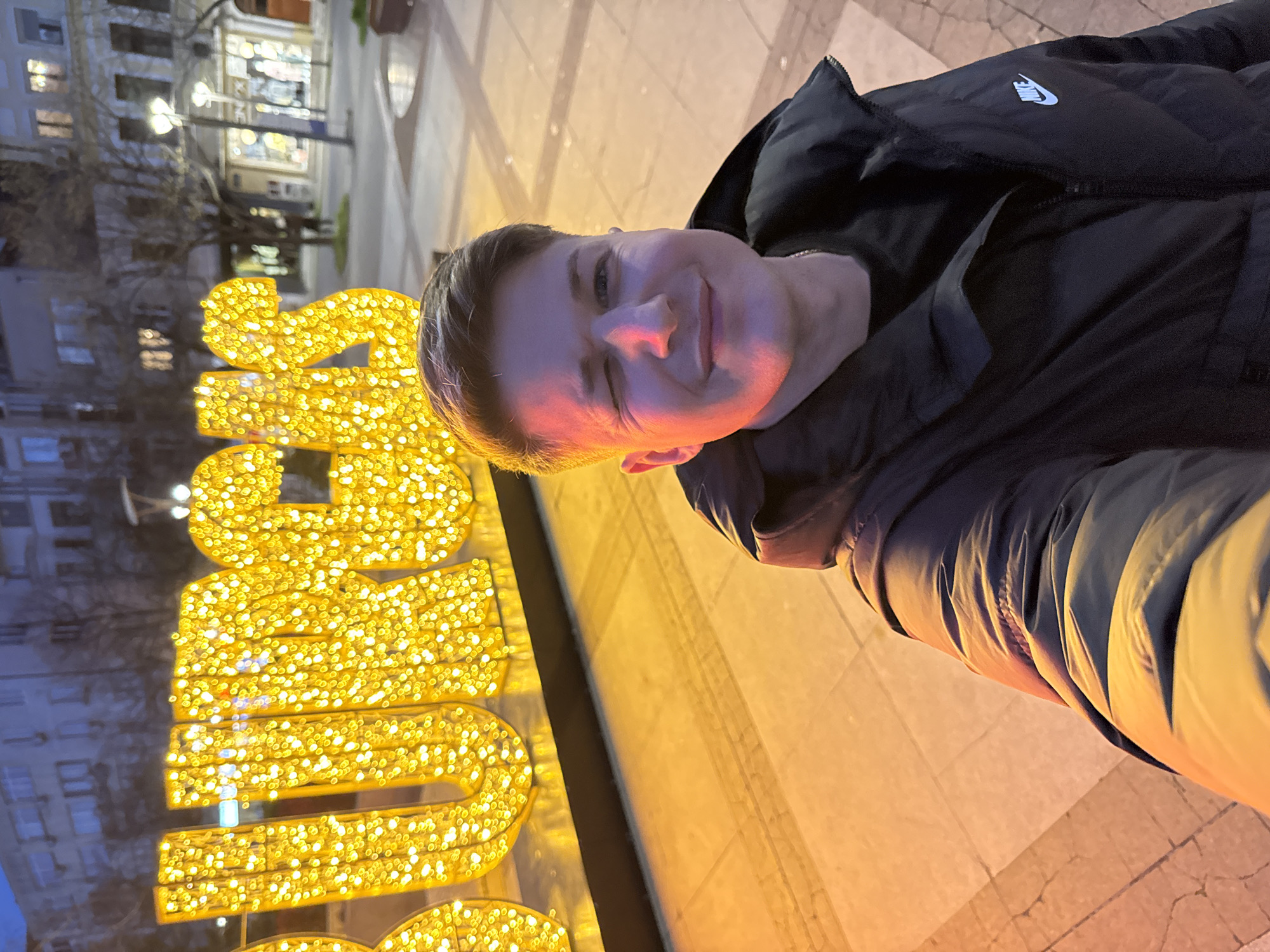 A rotated medium shot shows a person posing in front of a large, illuminated sign in Burgas, Bulgaria. The person, fair-skinned and wearing a black jacket with a Nike logo, is smiling slightly and looking towards the camera. The sign, composed of numerous small golden lights, is partially visible on the left side of the frame. The background shows a city street at night, with buildings and streetlights blurred in the distance. The ground is paved with light-colored tiles. The lighting suggests an evening setting, with a mix of artificial lights and some ambient light from the city.